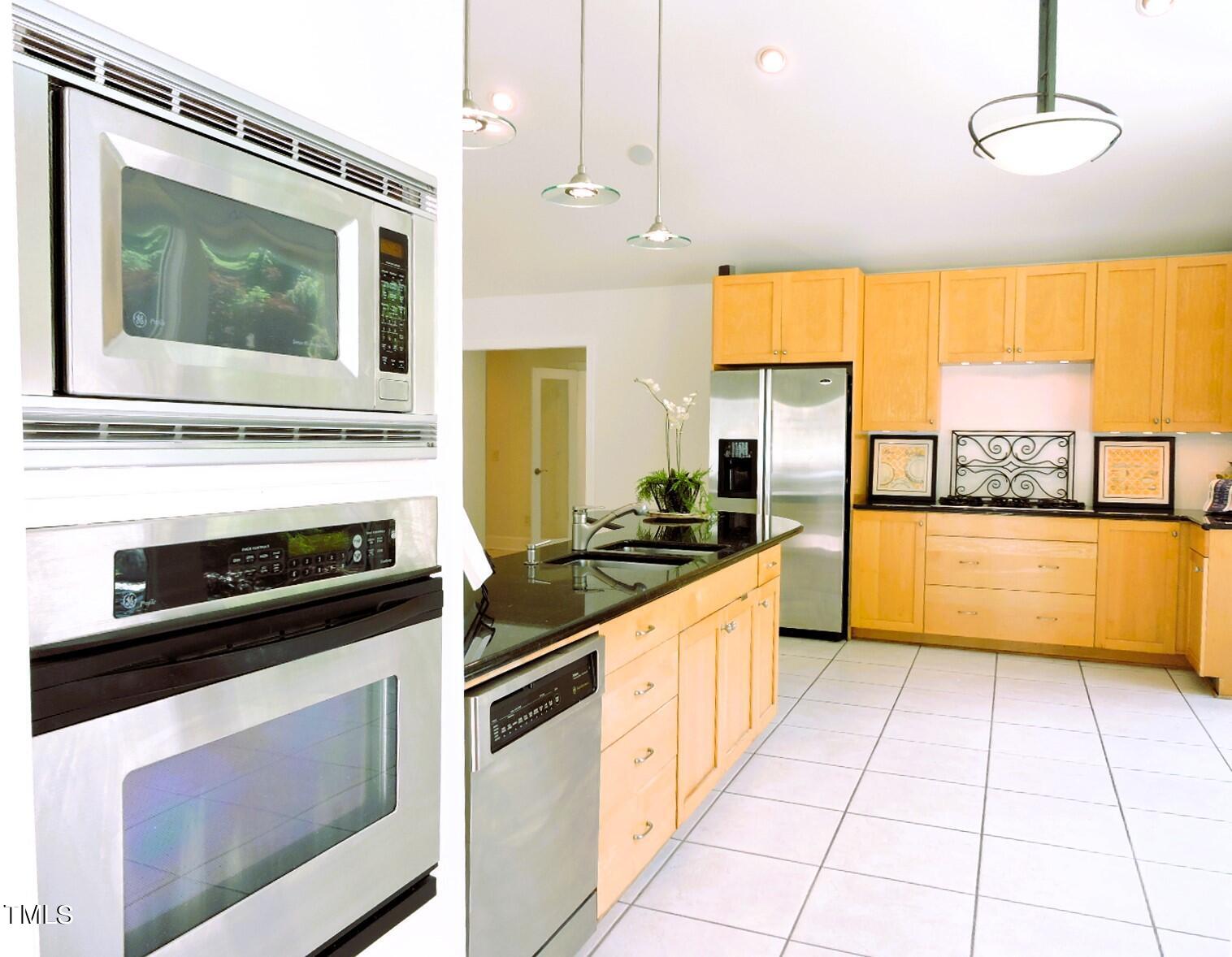Bright kitchen with maple cabinetry, stainless-steel appliances, dark counters, and a tiled floor.