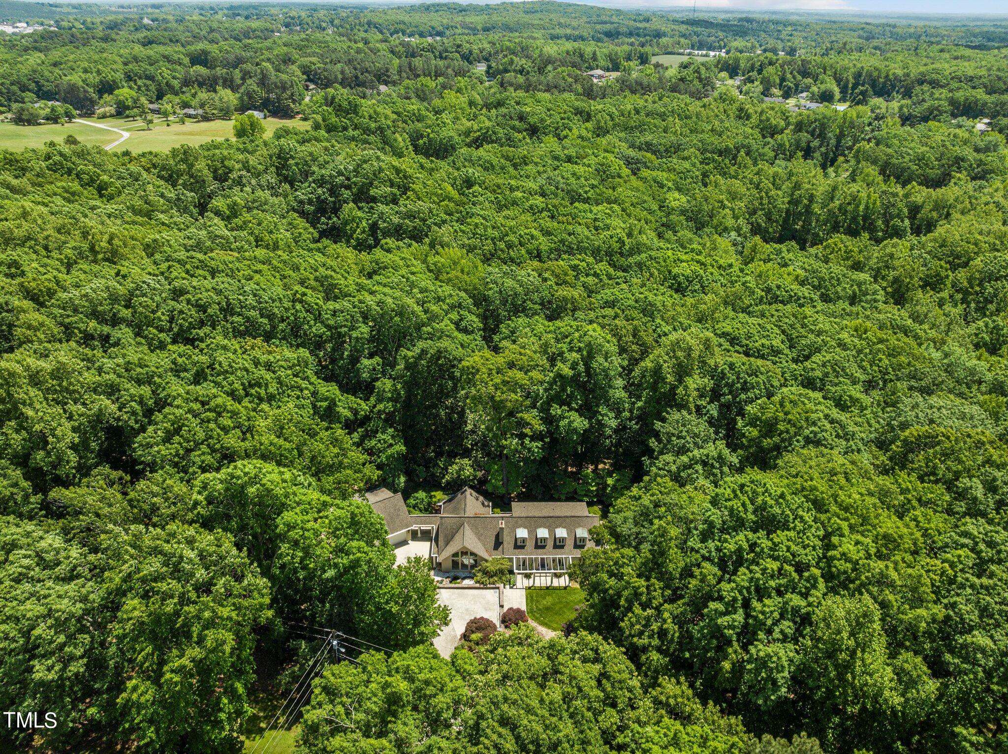 Aerial view of a house surrounded by dense woodland, with a driveway and small landscaped lawn.