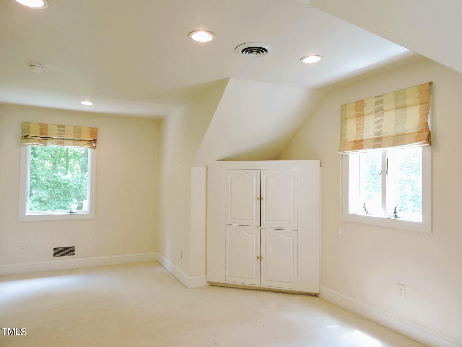 Vacant carpeted room with sloped ceiling, two windows, and built-in white cabinets.