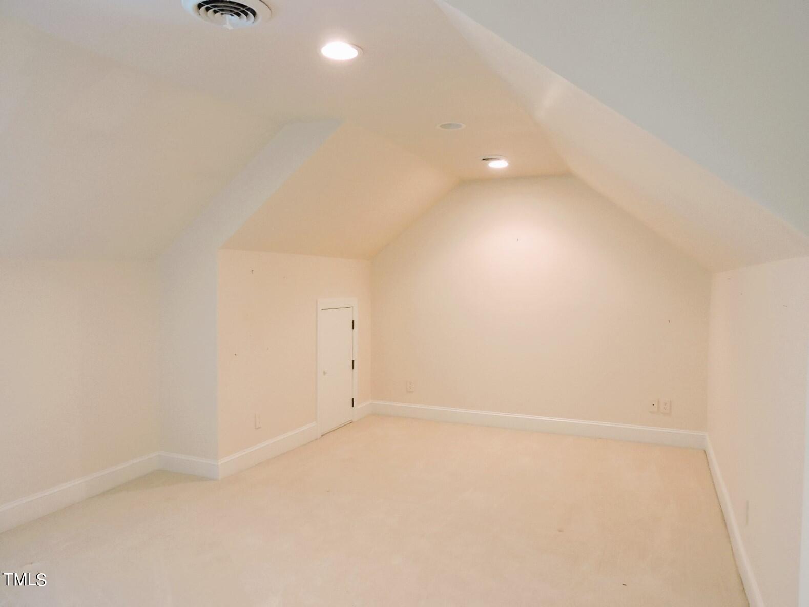 Empty carpeted room with a vaulted ceiling, recessed lights, and a small door.