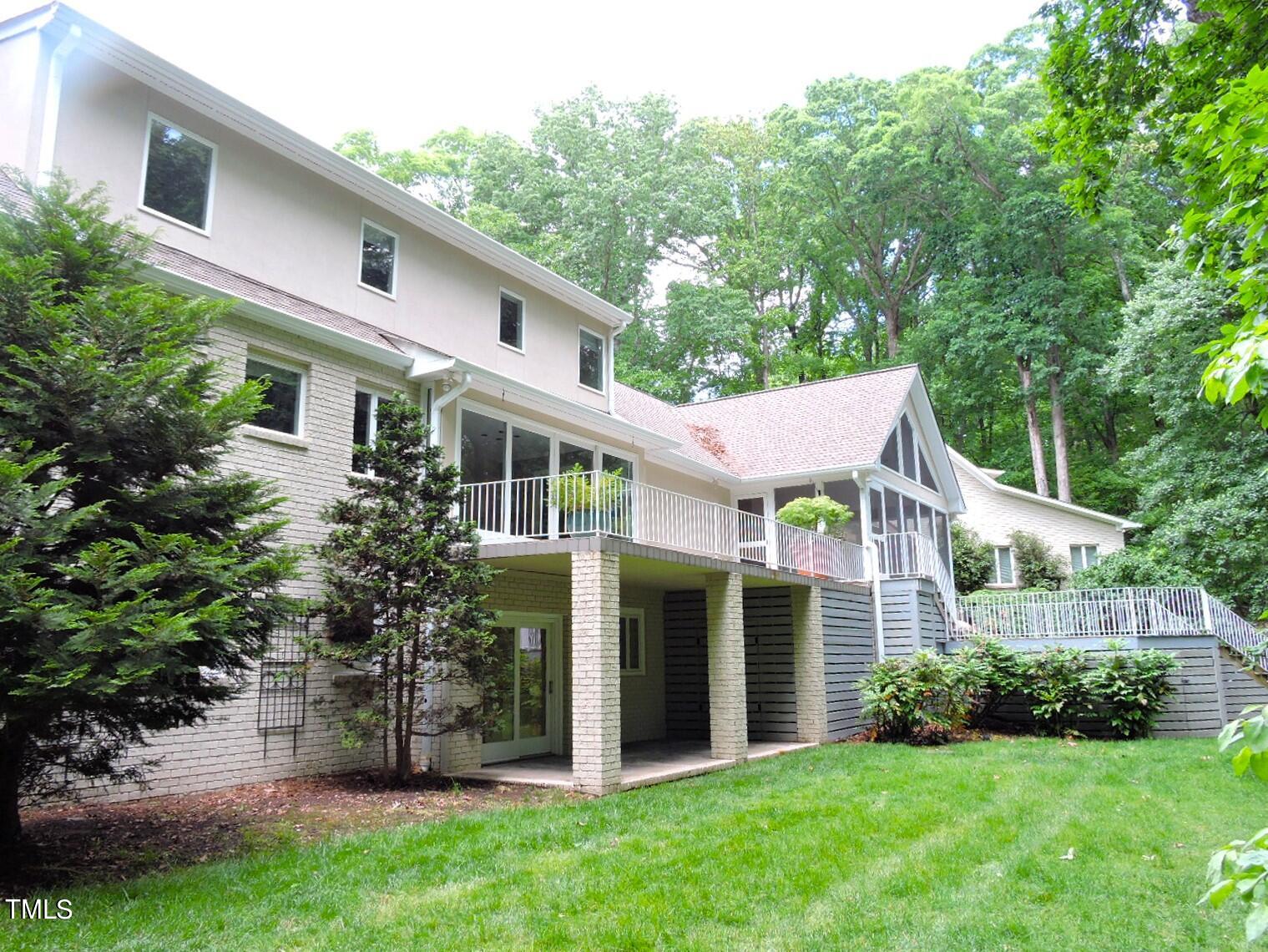 Rear of a light-colored house with a raised deck and screened porch amid leafy trees.