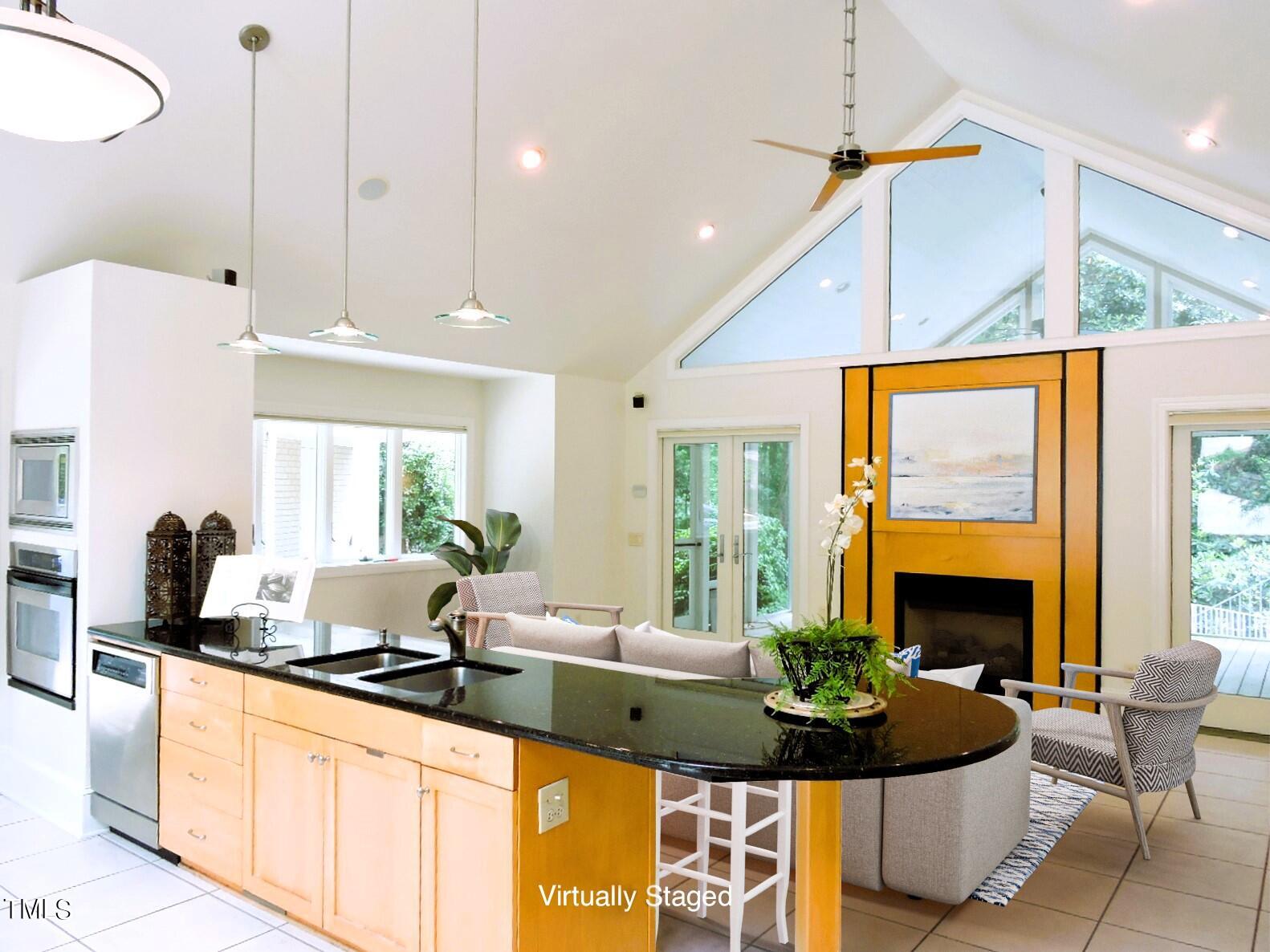 Open kitchen with a black island, light wood cabinets, vaulted ceiling, and virtually staged living area.