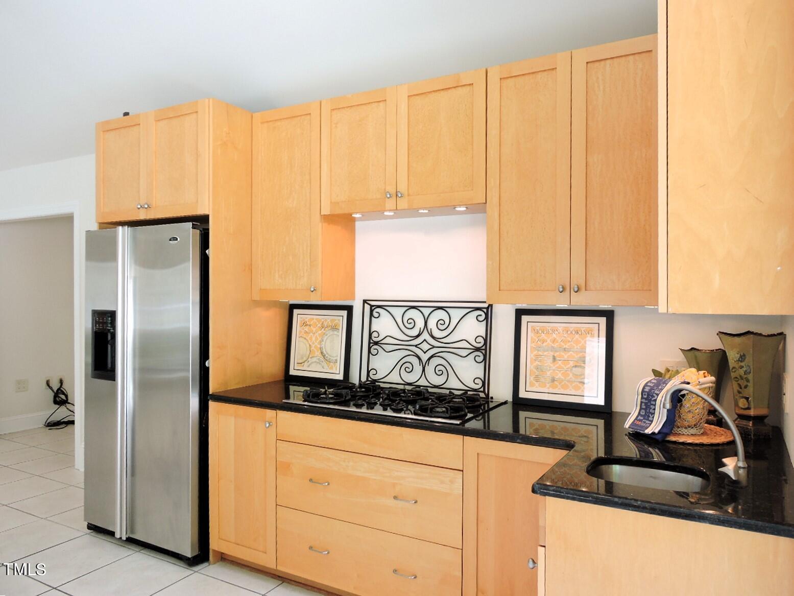 Kitchen with light wood cabinets, black countertops, gas cooktop, and stainless steel refrigerator.
