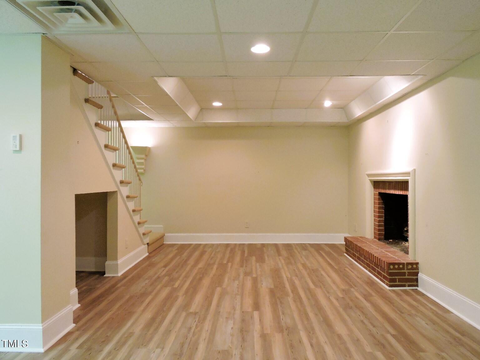 Empty basement room with wood-look flooring, staircase, and brick fireplace.
