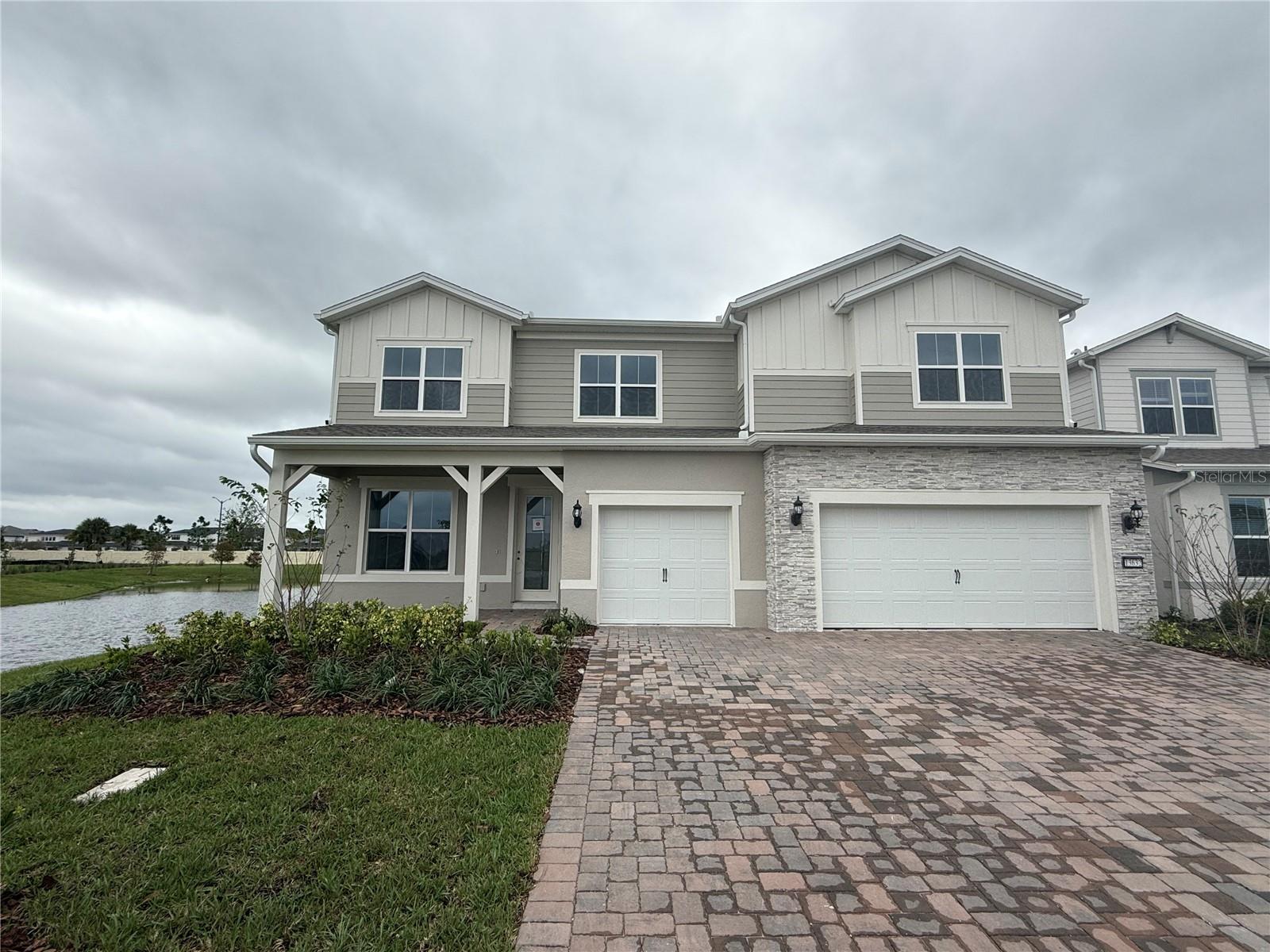 Two-story beige house with stone accents, a two-car garage, and a pond beside the lawn.