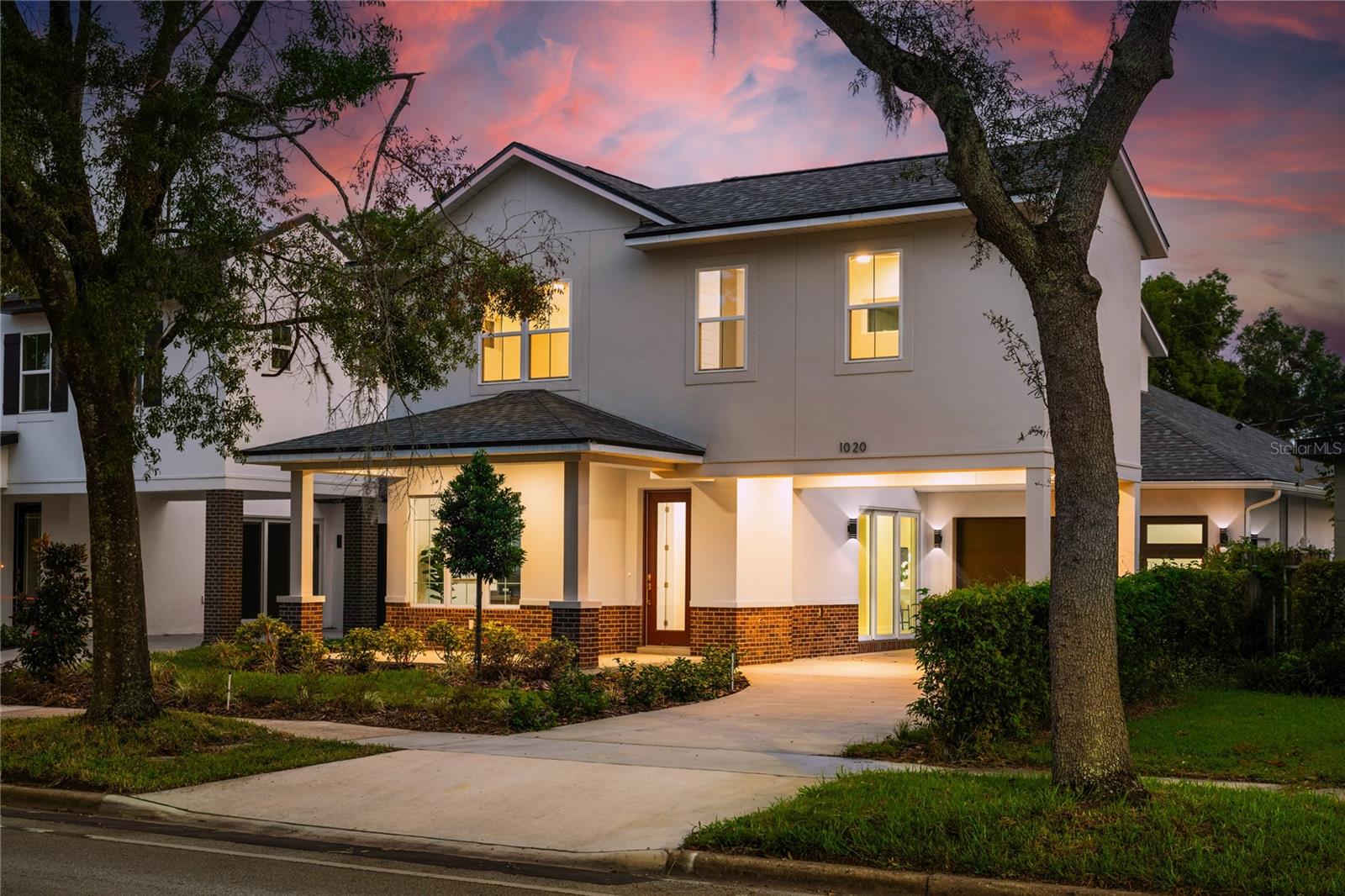 Two-story white house with brick accents and glowing windows, framed by trees at dusk.