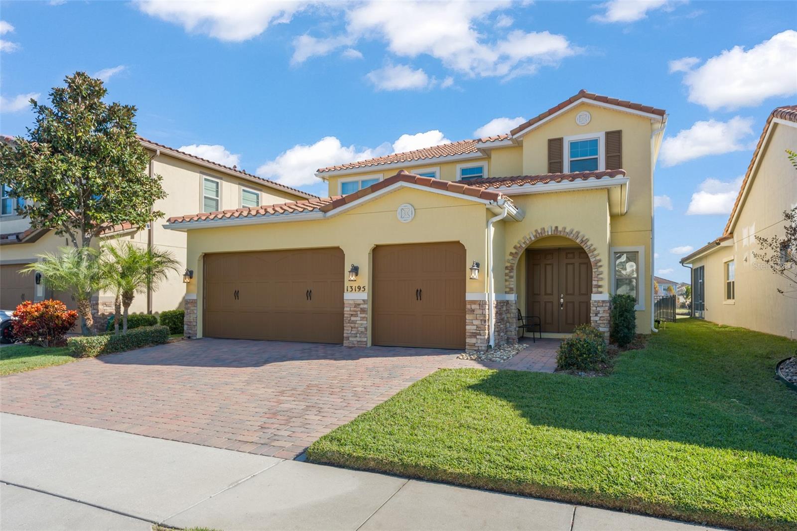 Yellow two-story home with terracotta roof, twin garage doors, arched entry, and paver driveway.