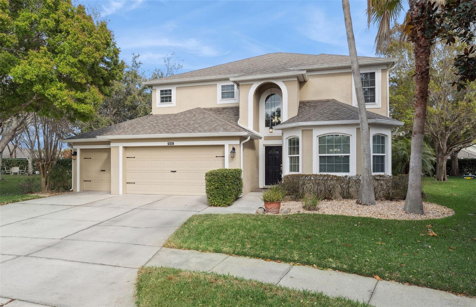 Two-story beige stucco home with an arched entry, attached garage, palm trees, and broad driveway.