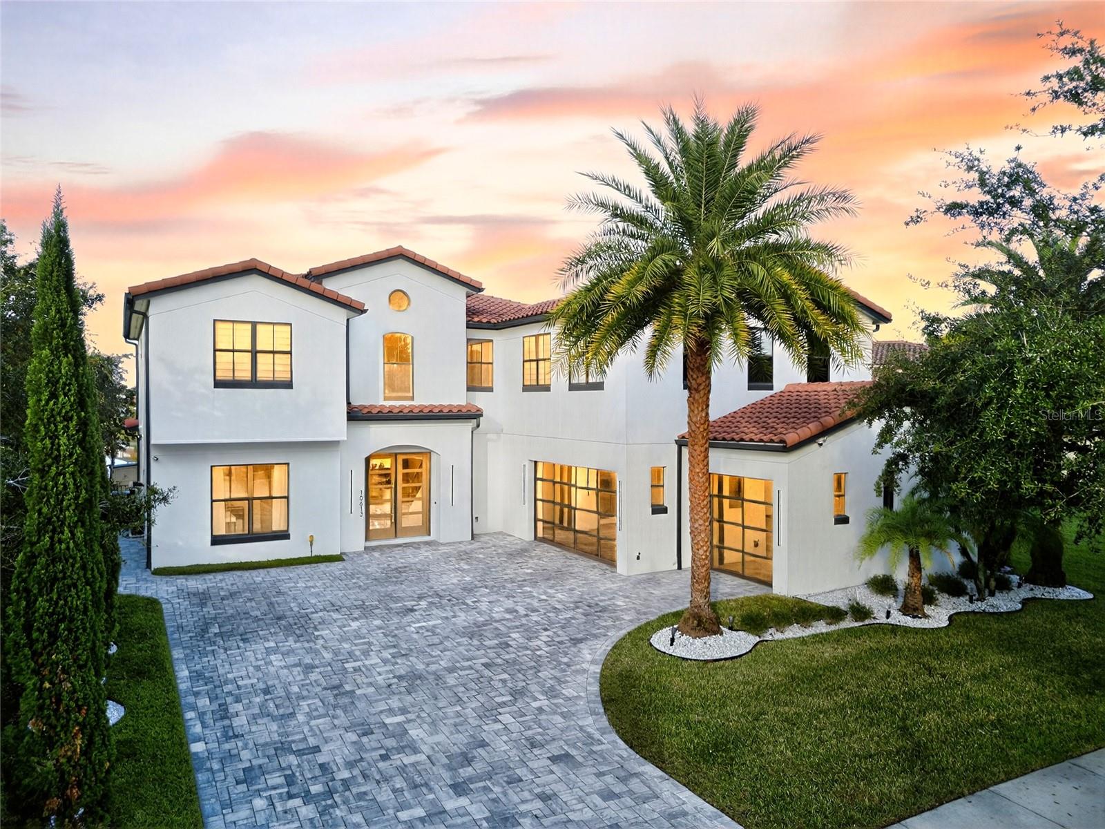White two-story house with a red tile roof, palm tree, and broad paved driveway at sunset.