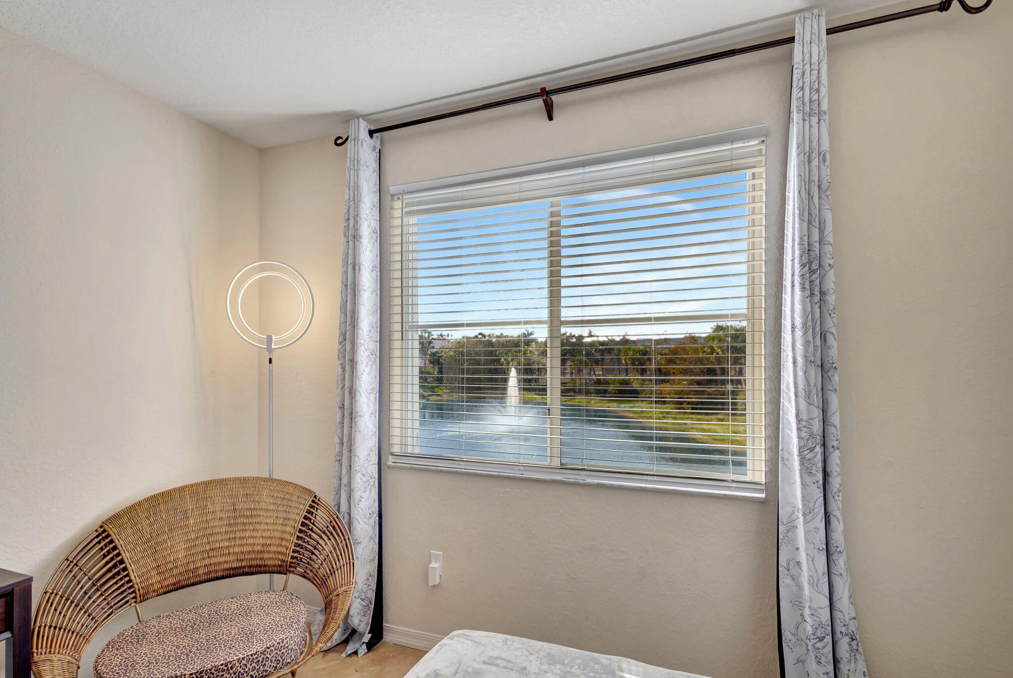 Bedroom corner with a wicker chair beside a window overlooking a pond and fountain.