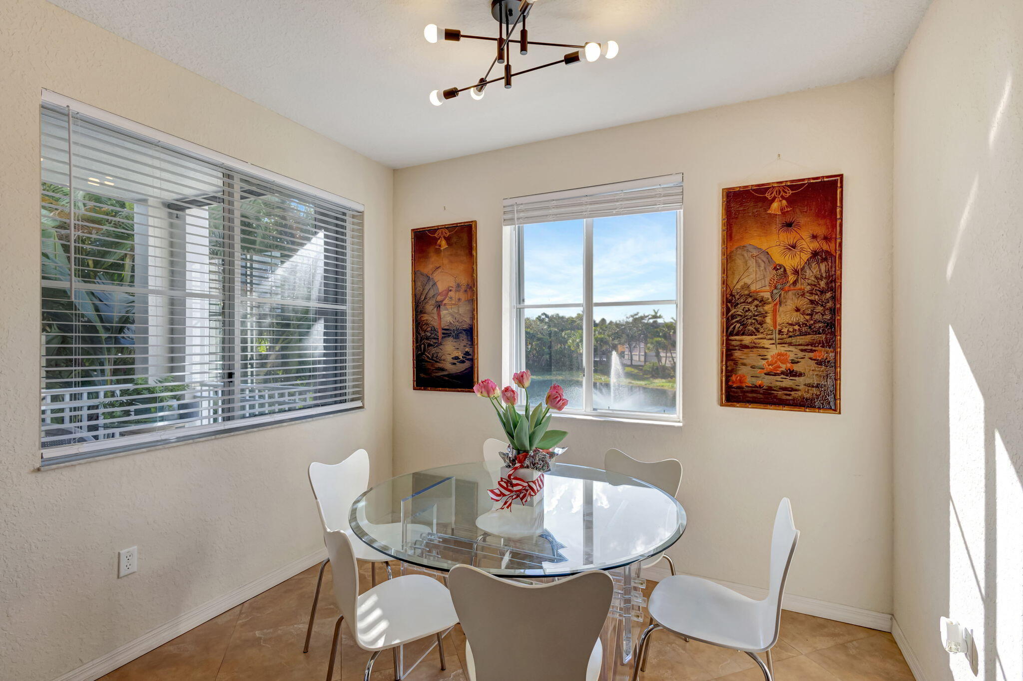 Dining area with a glass table, white chairs, and windows overlooking a pond fountain.