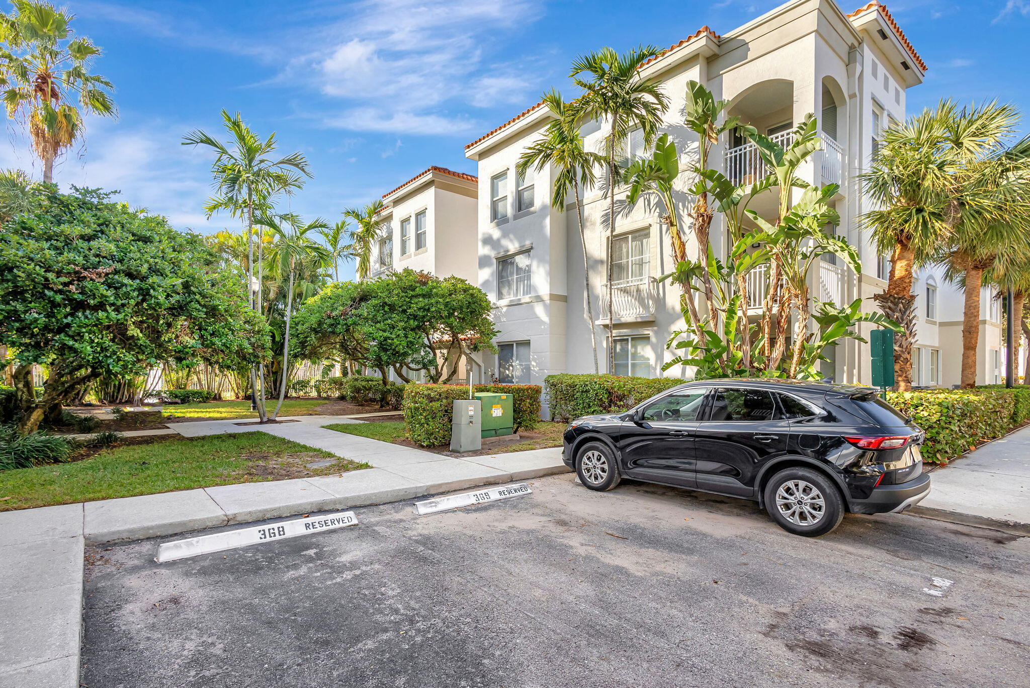 Cream stucco condominium building with arched balconies, tropical palms, and a black SUV parked out front.