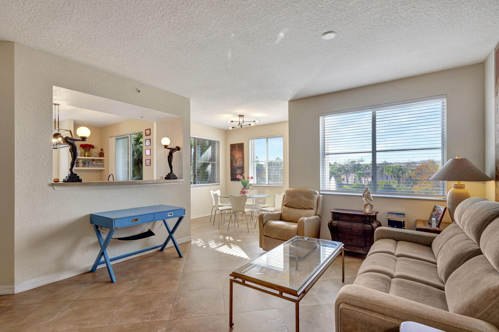 Living room with beige seating, a glass coffee table, and a bright window overlooking greenery.