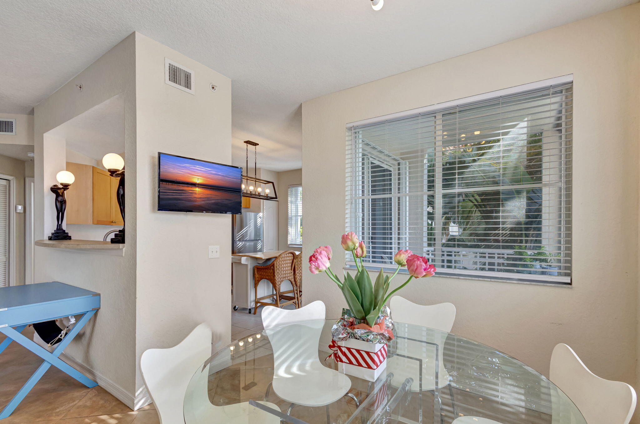 Dining area with a glass table, pink flowers, and a large window overlooking tropical greenery.