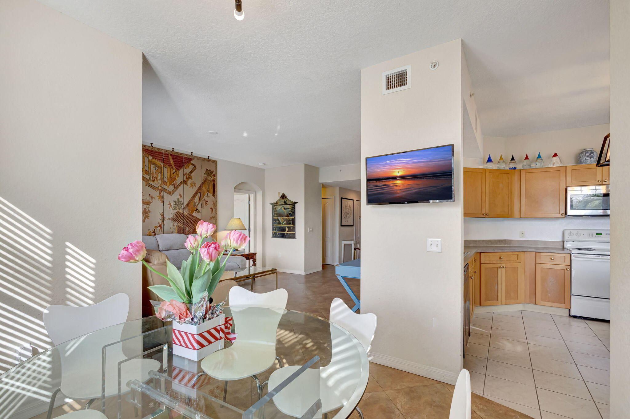Furnished condo dining area and kitchen, with pink flowers on a glass table and a sunset displayed on the television.