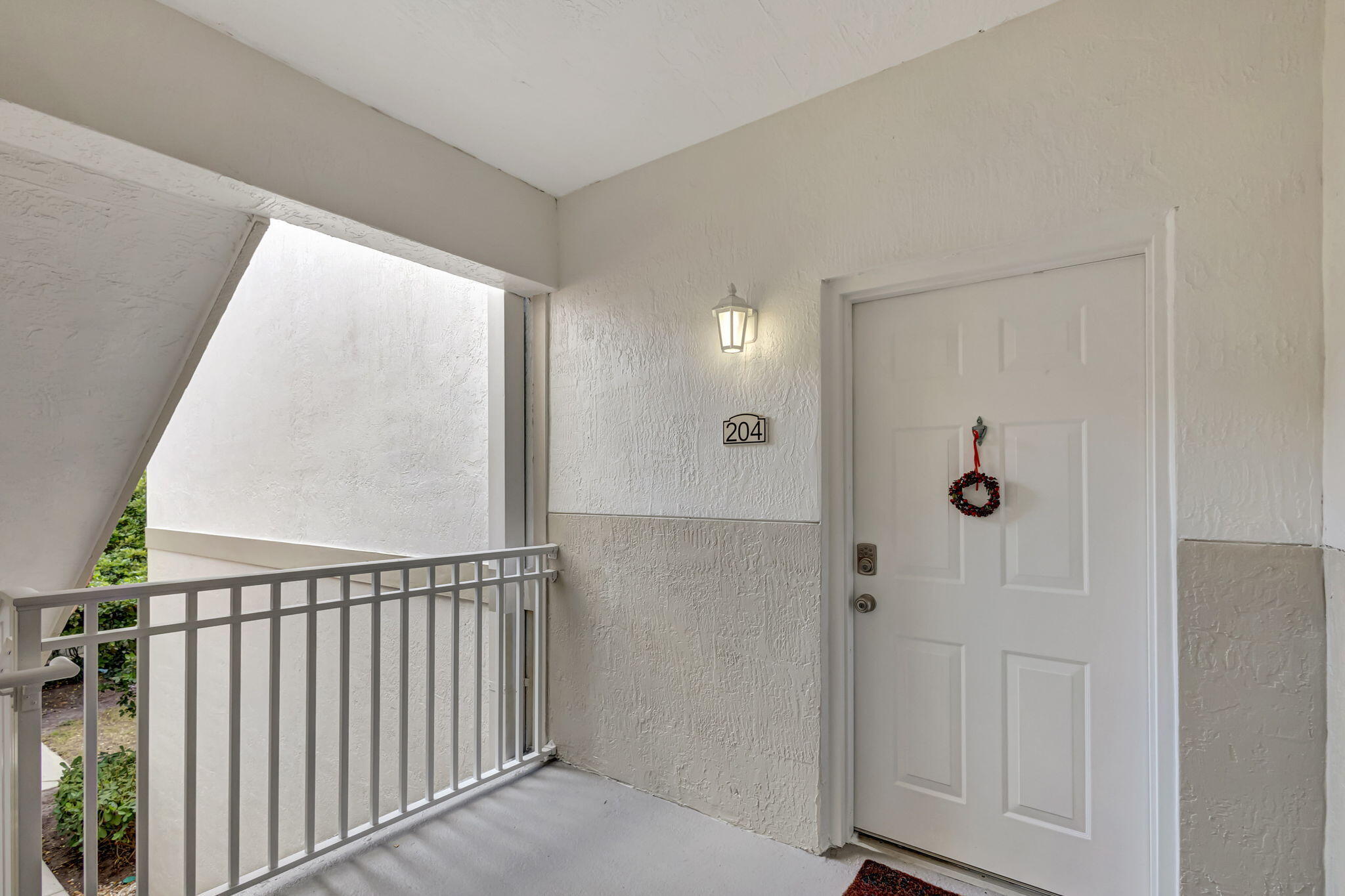 Covered condo entry with unit 204 plaque, white front door, wreath, and metal walkway railing.