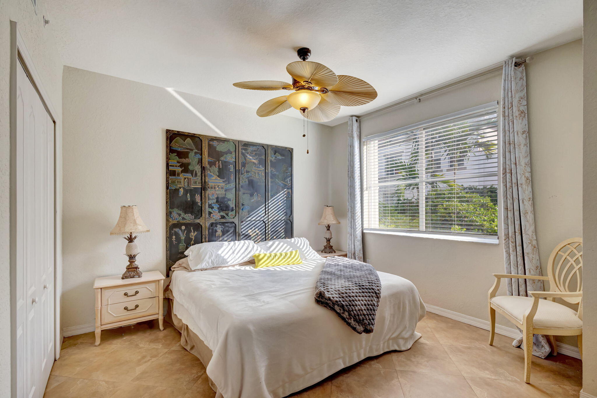 Bedroom with a white bed, decorative folding screen, ceiling fan, and sunlit window overlooking greenery.