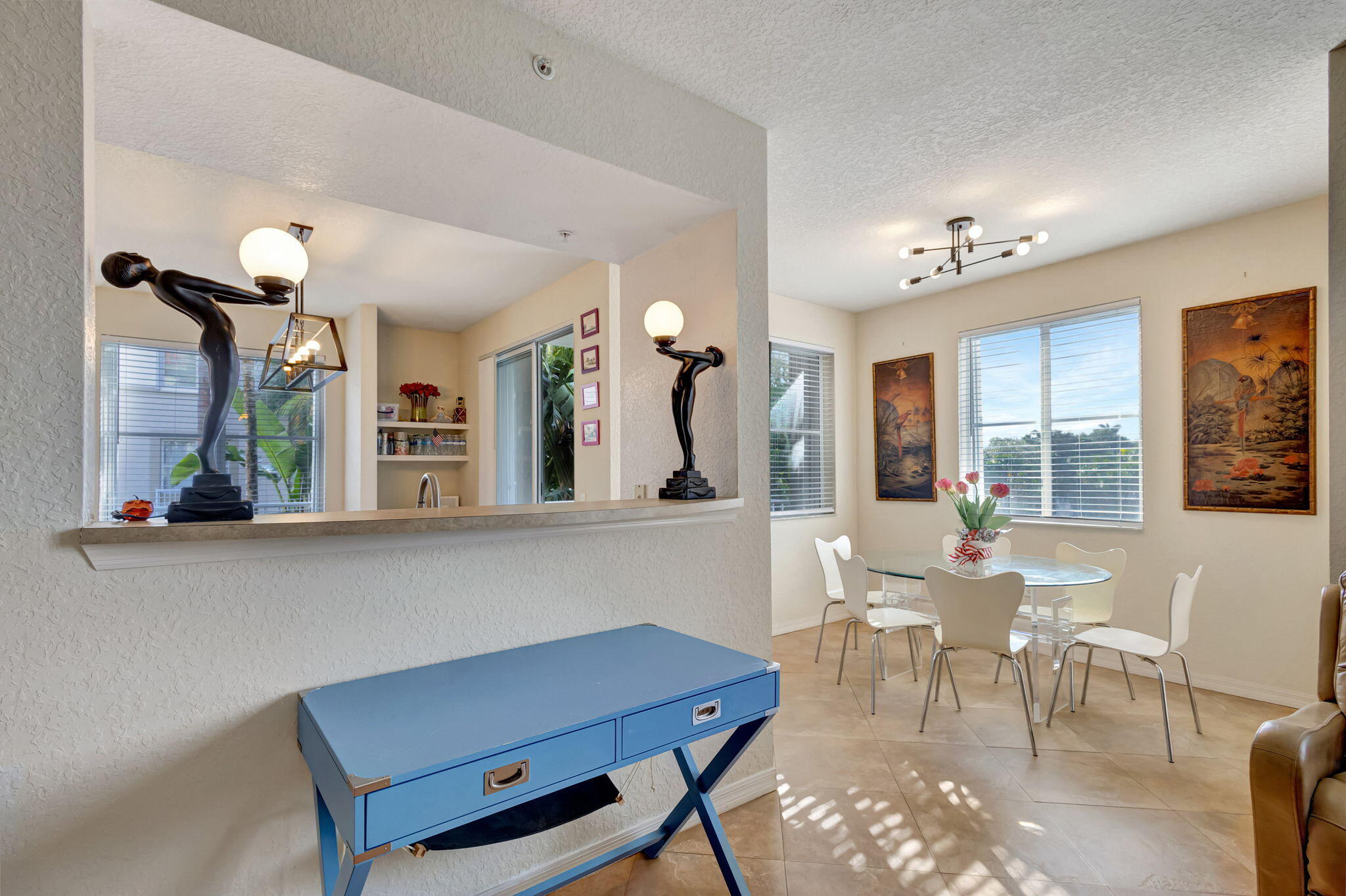 Dining area with a glass table, white chairs, and sunlit windows beside a pass-through.