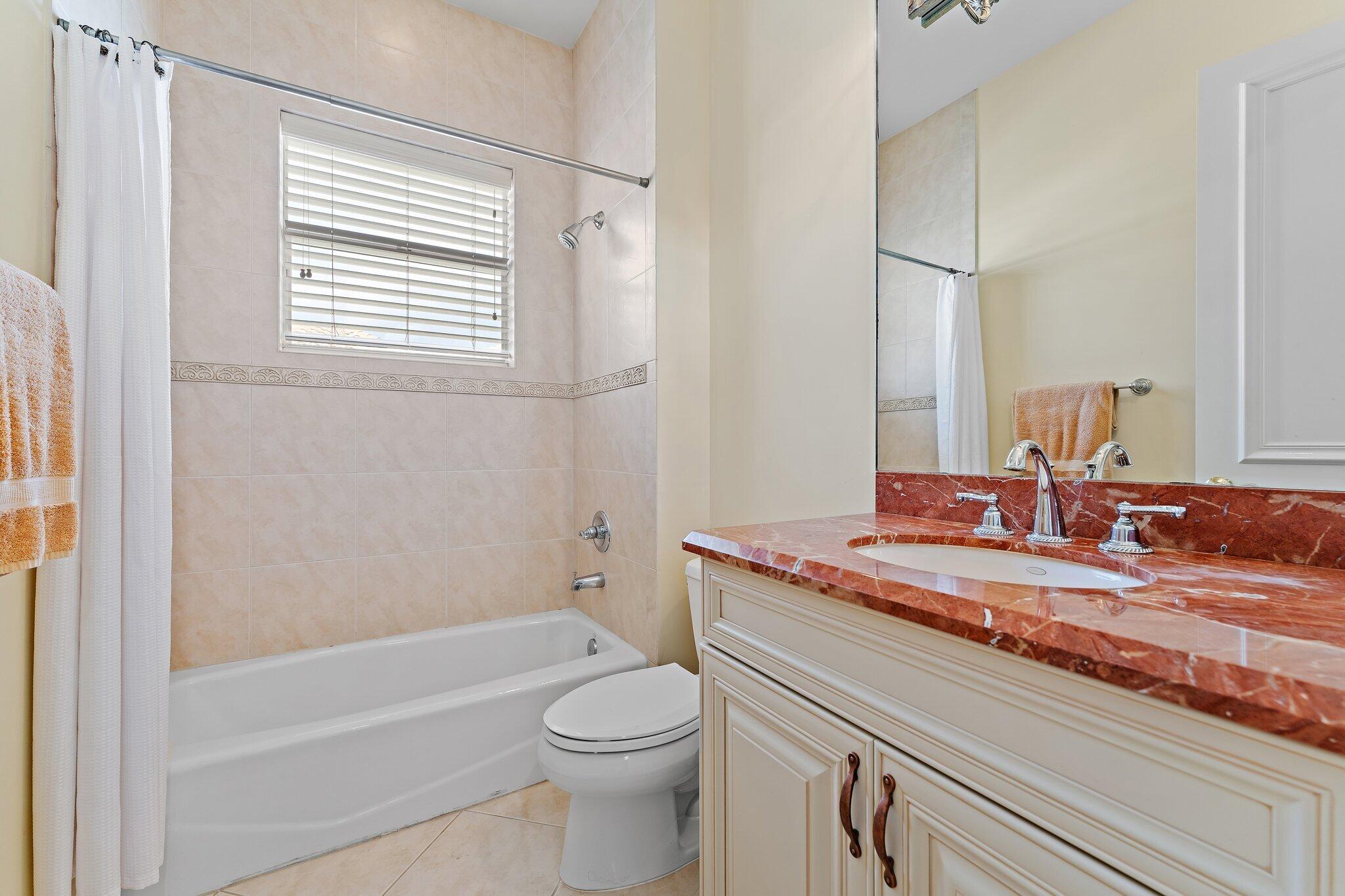 Bathroom with a white bathtub, window blinds, and cream vanity with a reddish marble countertop.