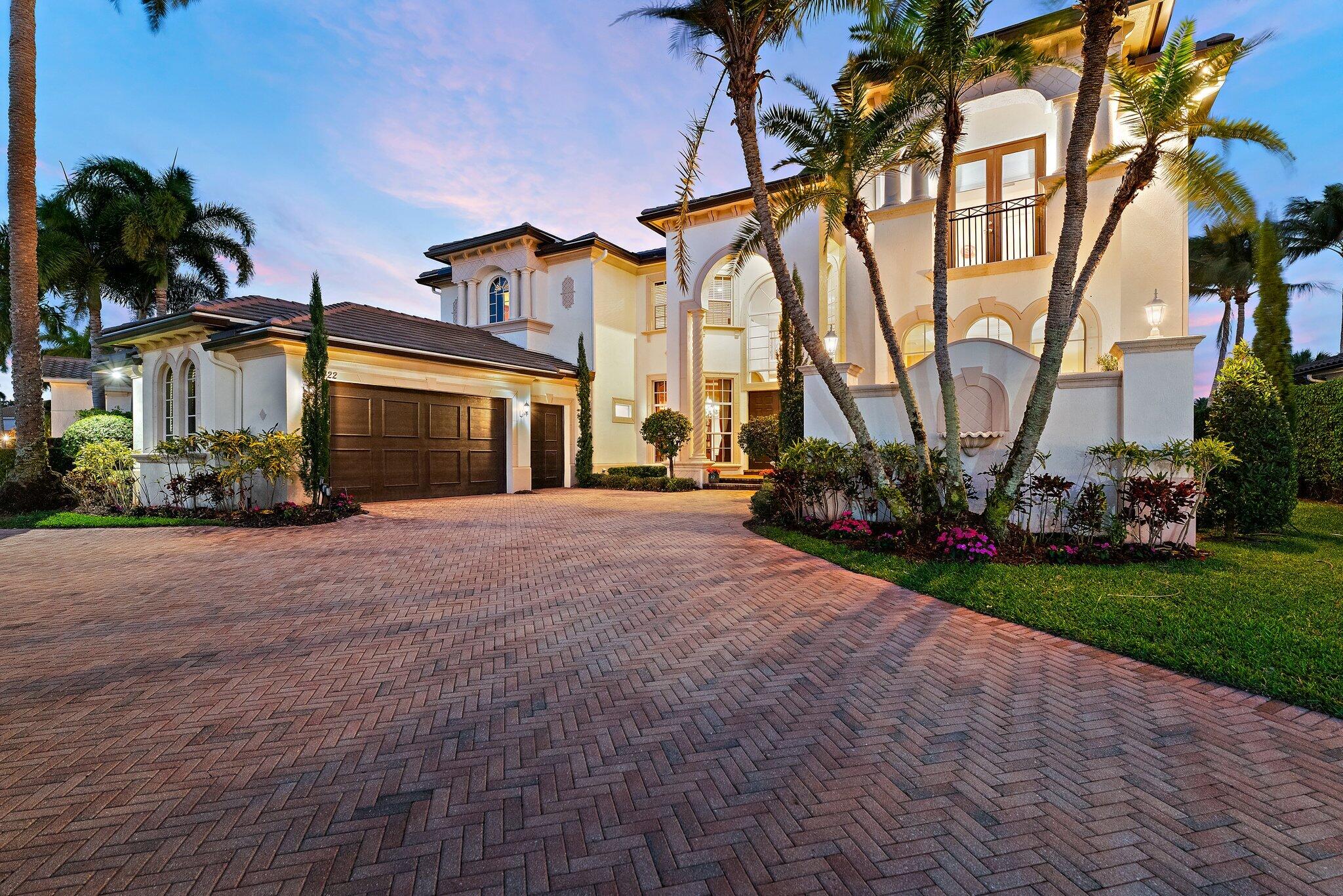 Mediterranean-style home with arched windows, palm trees, and a broad brick driveway at dusk.