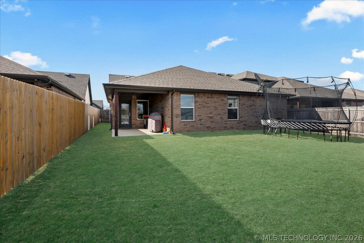 Brick home’s backyard with a covered patio, large lawn, wooden privacy fence, and trampoline.