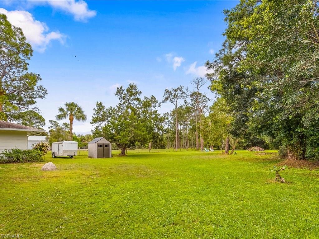 Spacious grassy yard framed by mature trees, with a small shed and utility trailer.
