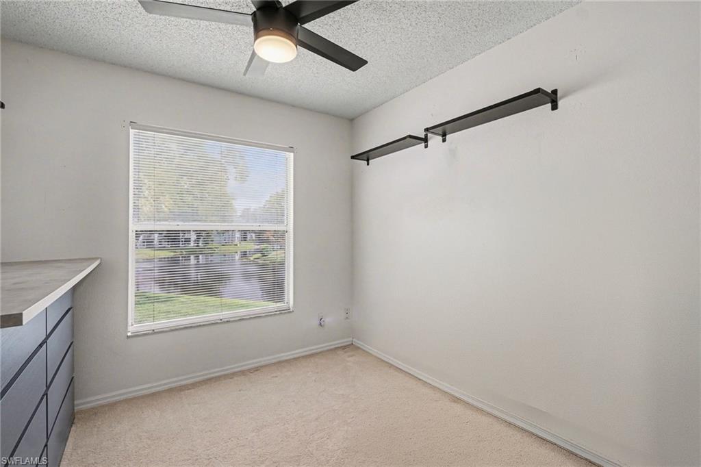 Empty bedroom with beige carpet, window blinds, wall shelves, and a ceiling fan.