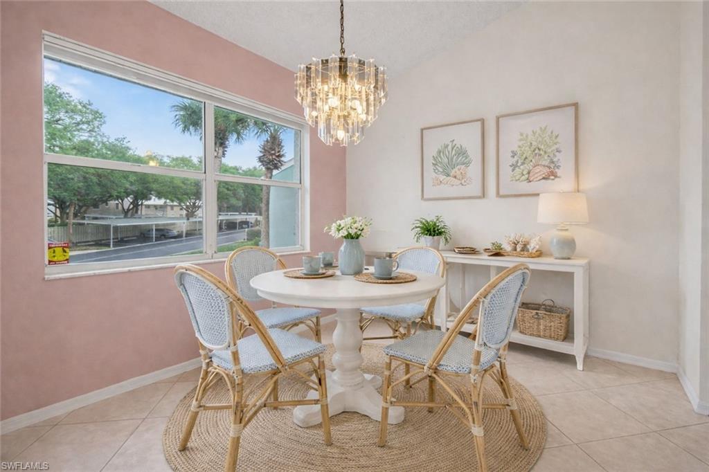Dining room with a round white table, woven chairs, and crystal chandelier beside large windows.
