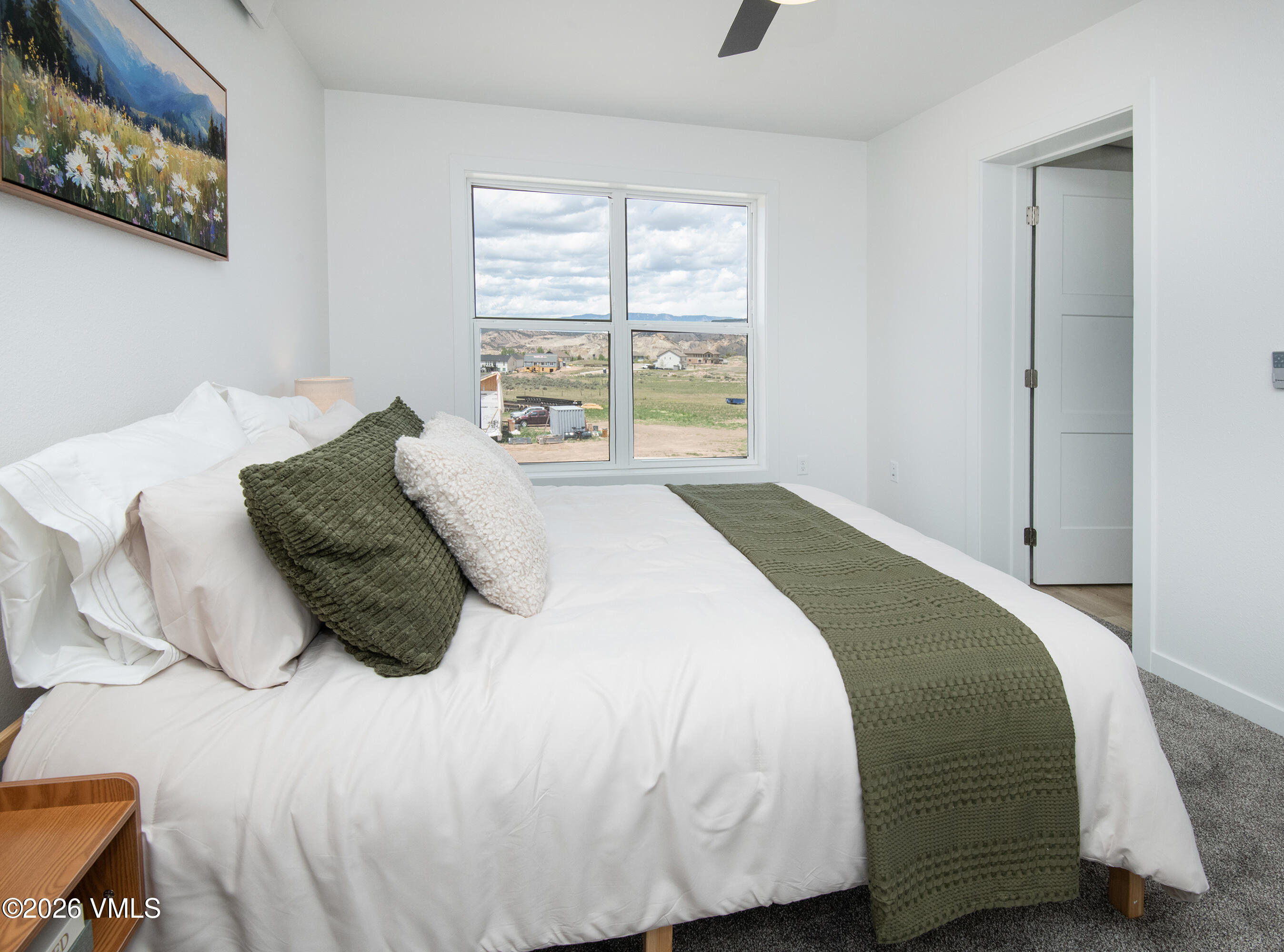 Staged bedroom with a white bed, green accents, and a window overlooking open fields and distant hills.