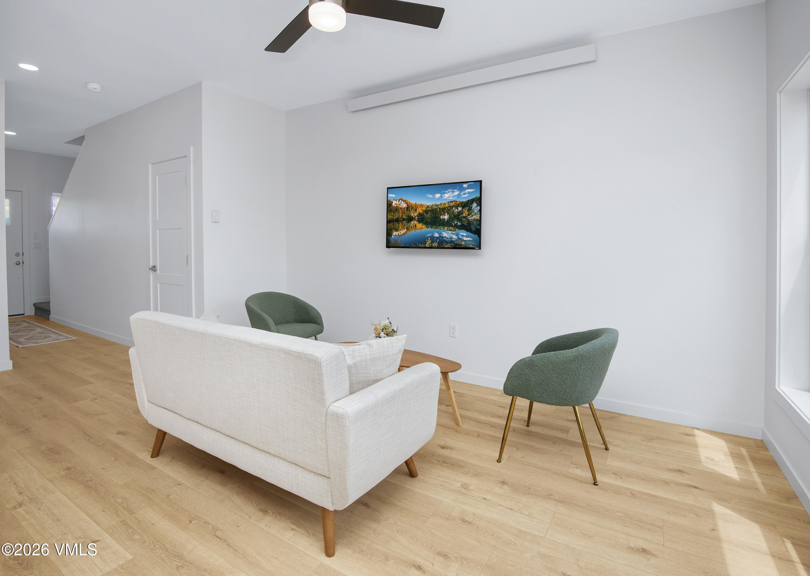 Furnished living room with a white sofa, green chairs, wood floors, and a wall-mounted television.