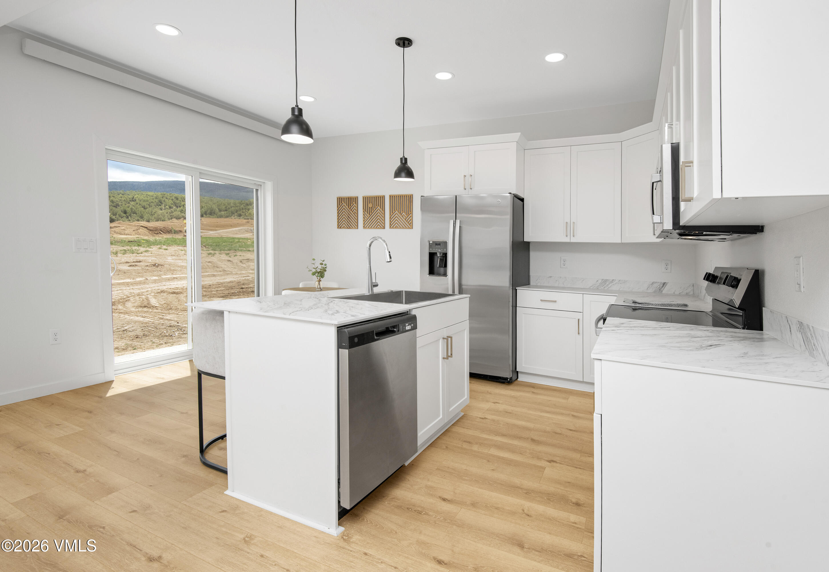 Modern white kitchen with stainless steel appliances, a central island, and wooded hills visible through sliding doors.