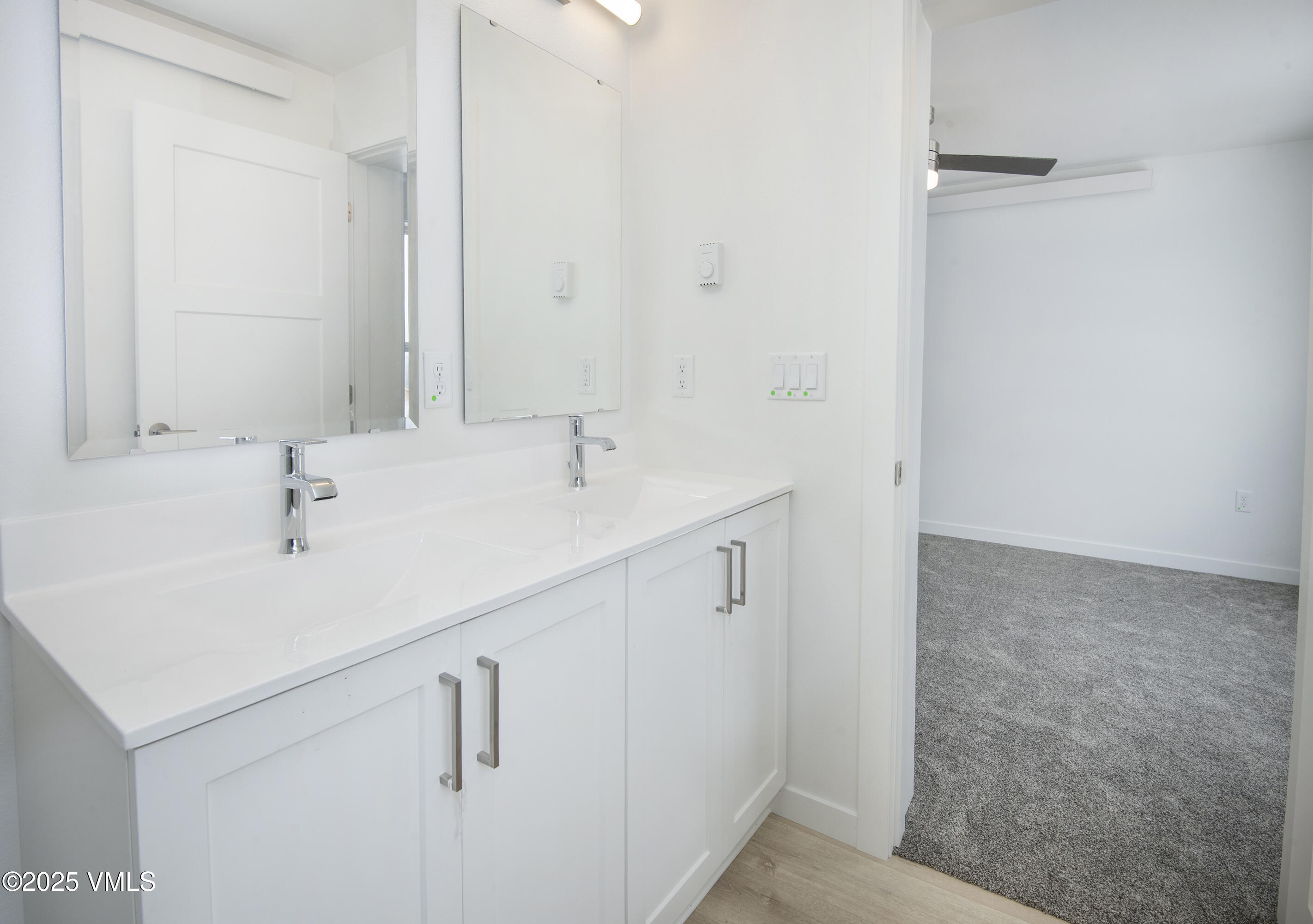 Bathroom with a white double-sink vanity and mirrored cabinets beside an open doorway to a carpeted room.