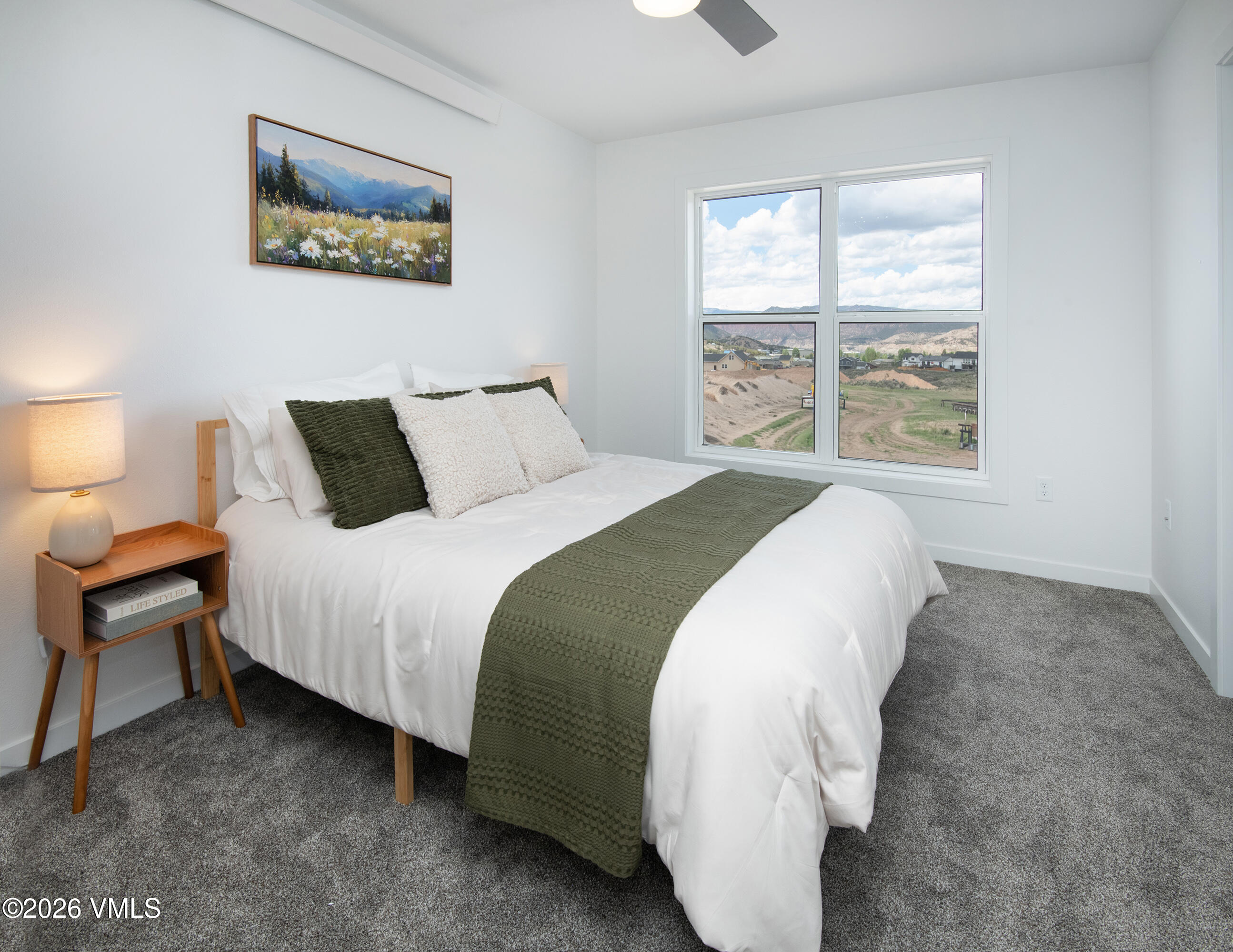 Staged bedroom with a white bed and green accents, looking toward distant hills through a large window.