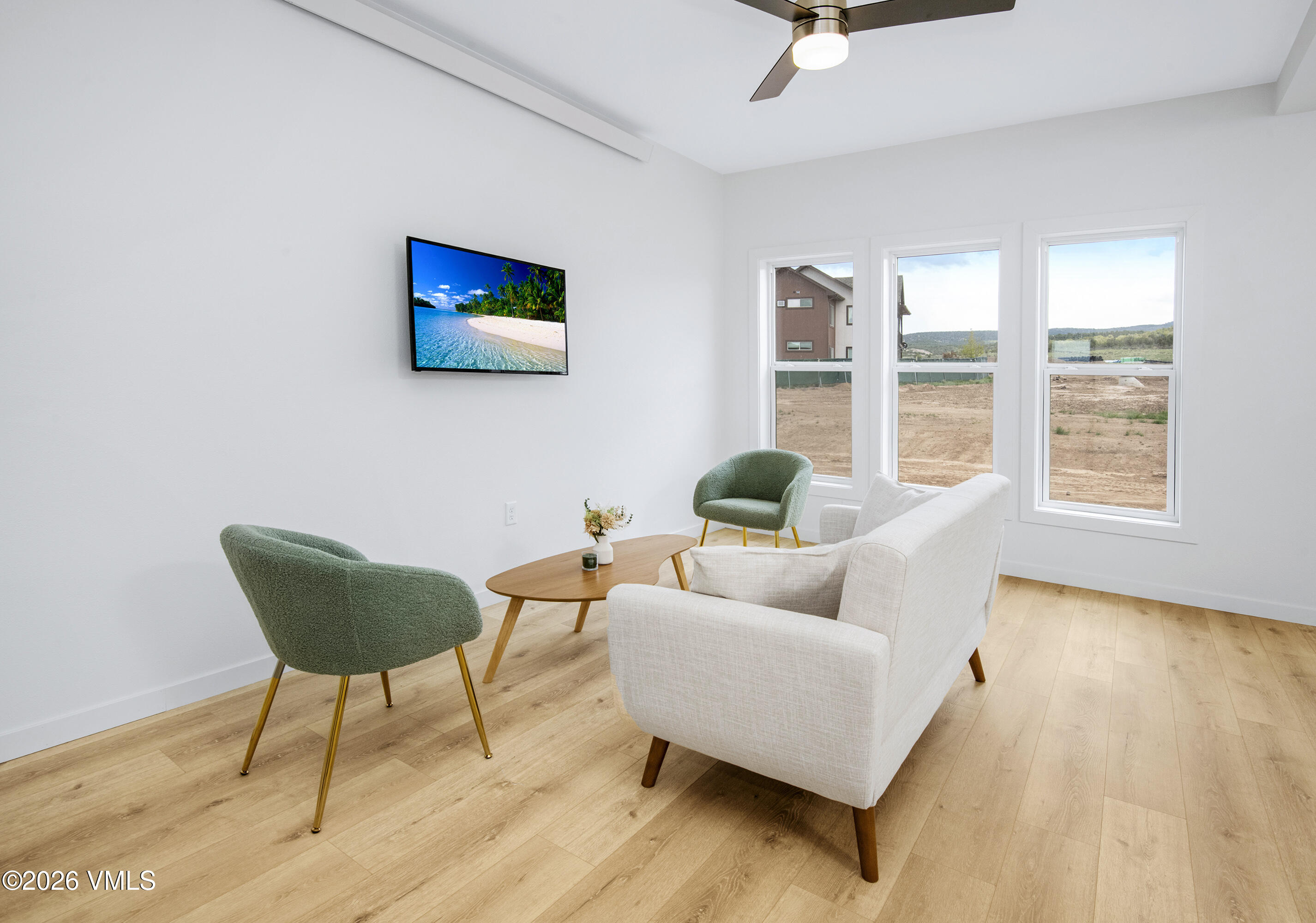Staged living room with green accent chairs, light wood floors, and three windows overlooking a bare yard.