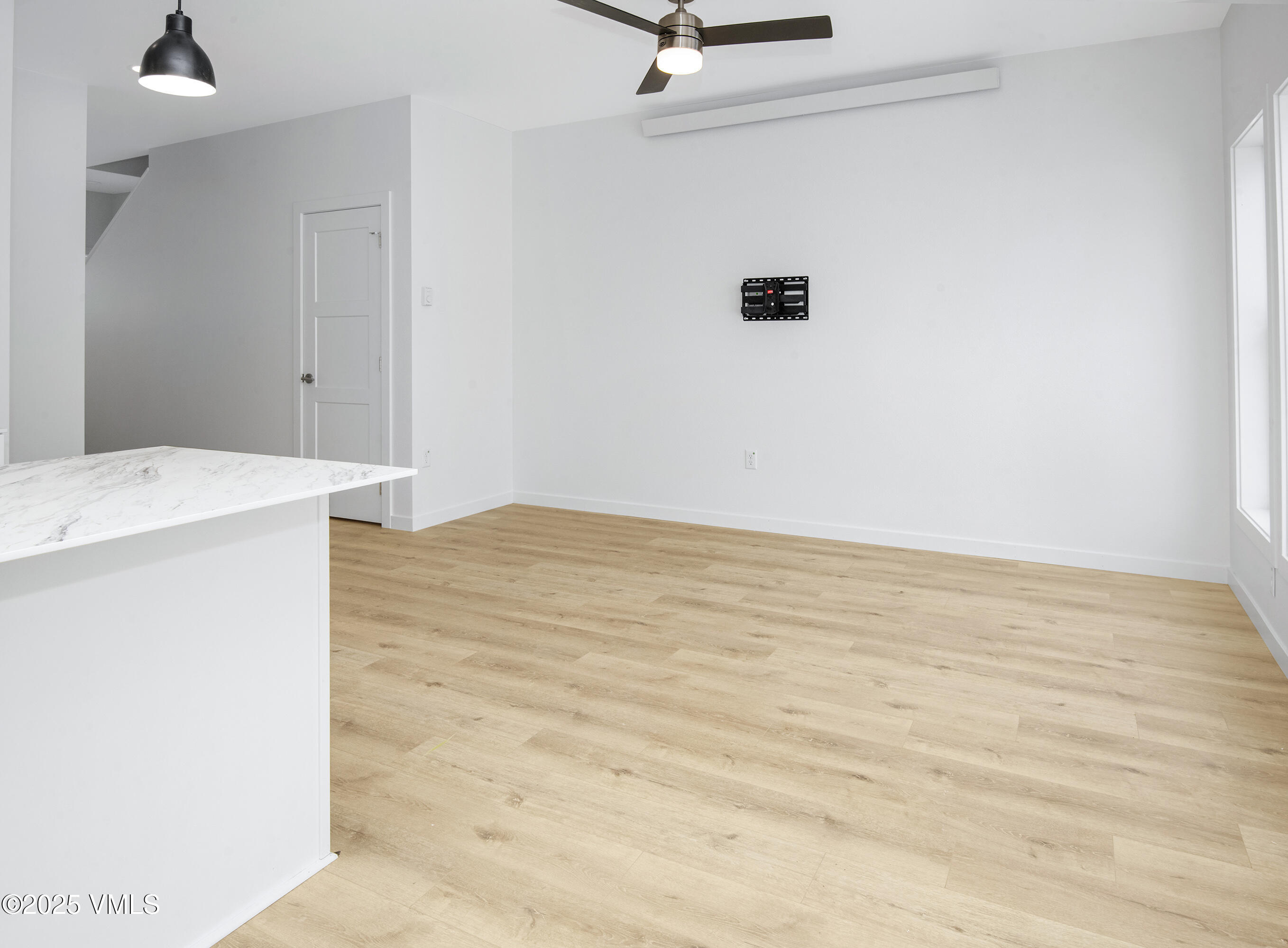 Vacant living area with light wood flooring, ceiling fan, TV bracket, and a marble-look island counter.