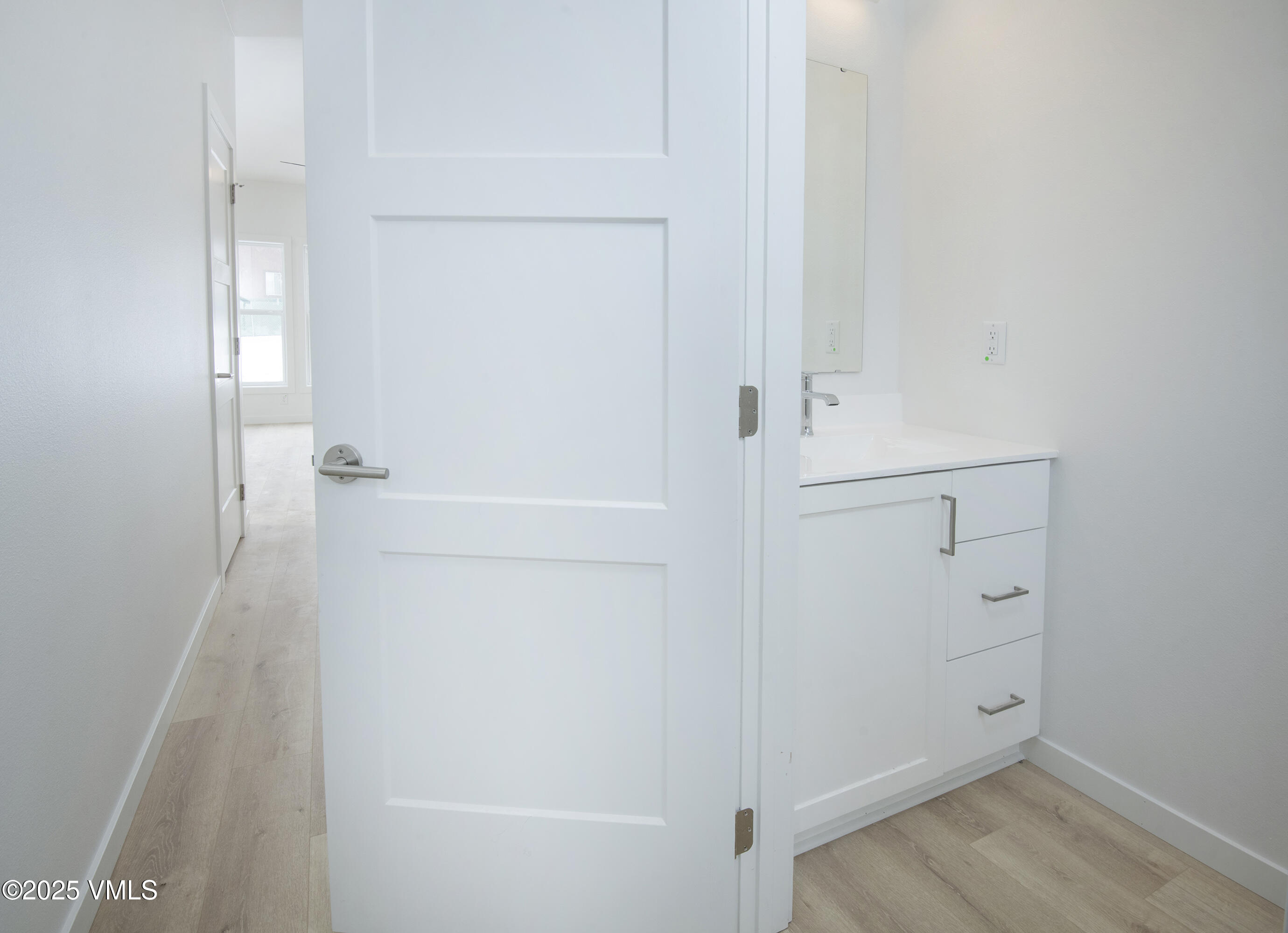 Bathroom vanity behind an open white door, with light wood-look flooring and a hallway beyond.