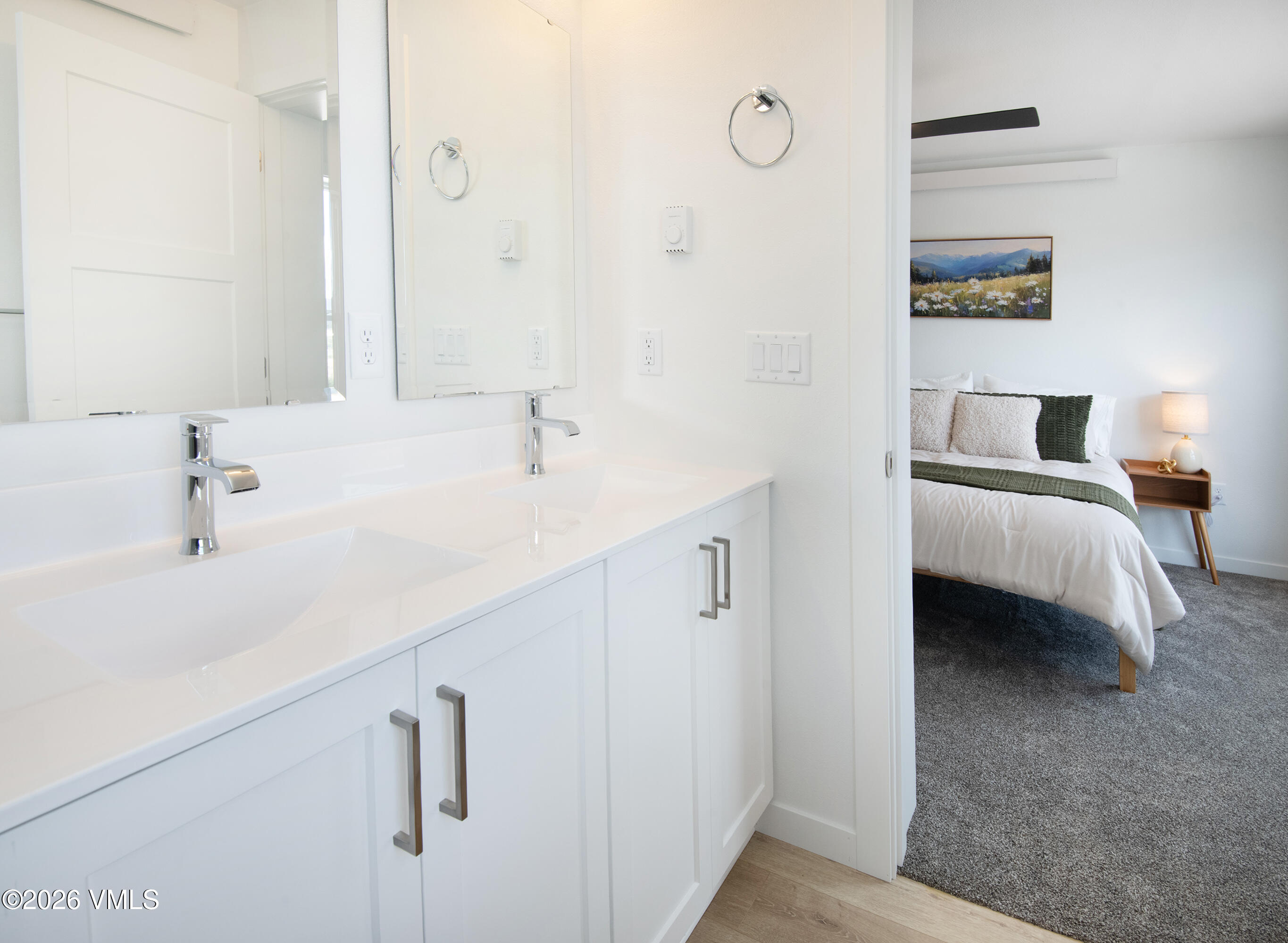 Double-sink bathroom vanity with large mirrors; a bedroom with green accents is visible through the doorway.