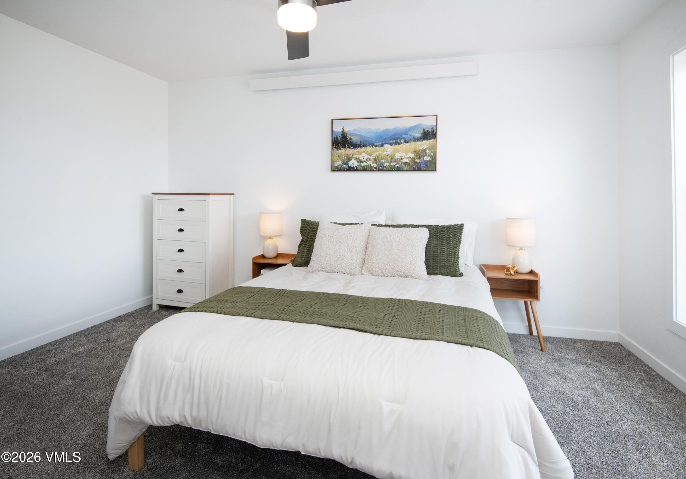 Staged bedroom with a white bed, olive-green accents, matching bedside lamps, and a mountain wildflower painting.