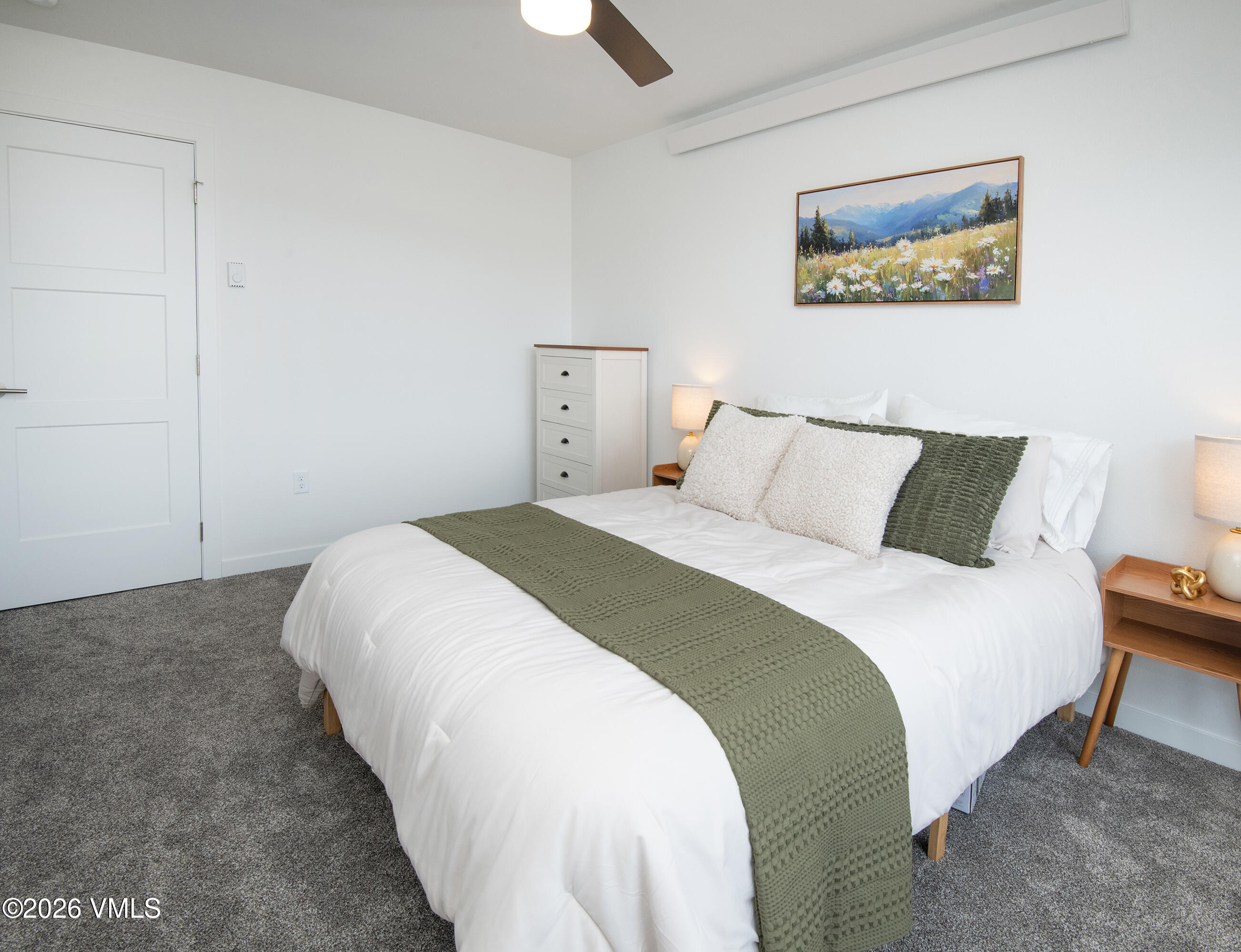 Bedroom with white bedding, an olive-green throw, gray carpet, and mountain-meadow artwork.