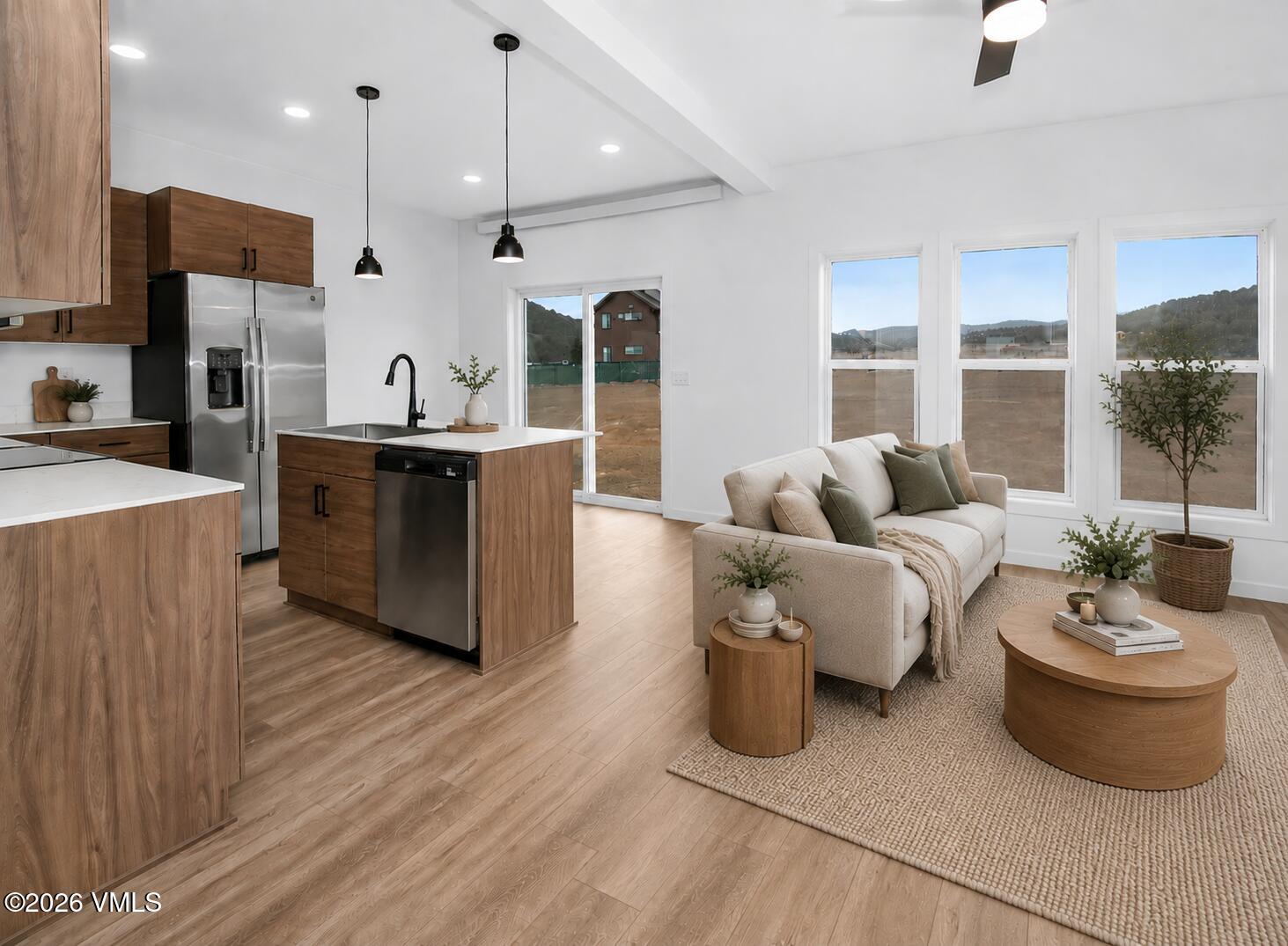 Open-plan kitchen and living room with wood cabinetry, stainless appliances, and a neutral sofa by tall windows.