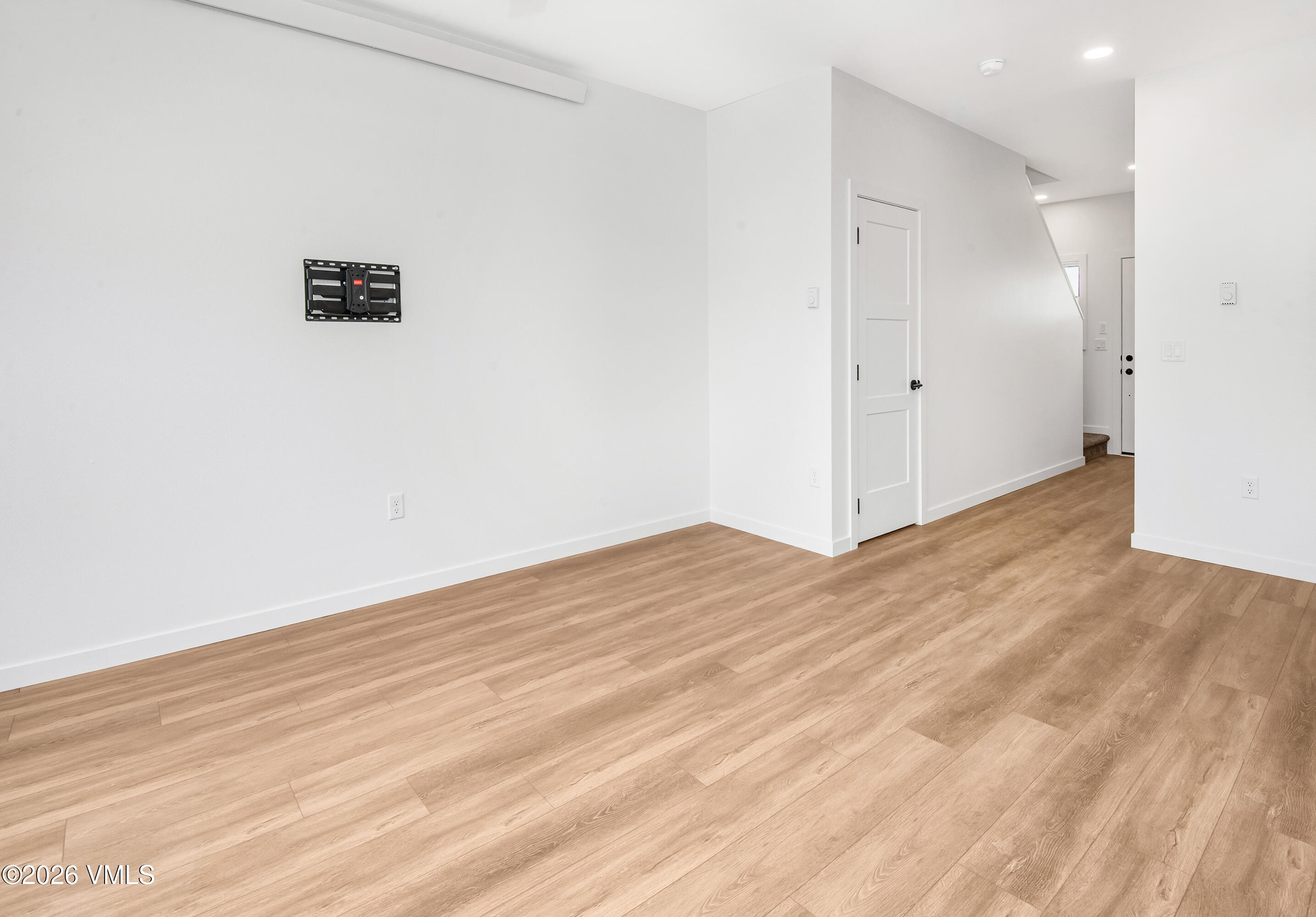 Empty room with light wood-look flooring, a wall-mounted TV bracket, and a hallway to the entry.
