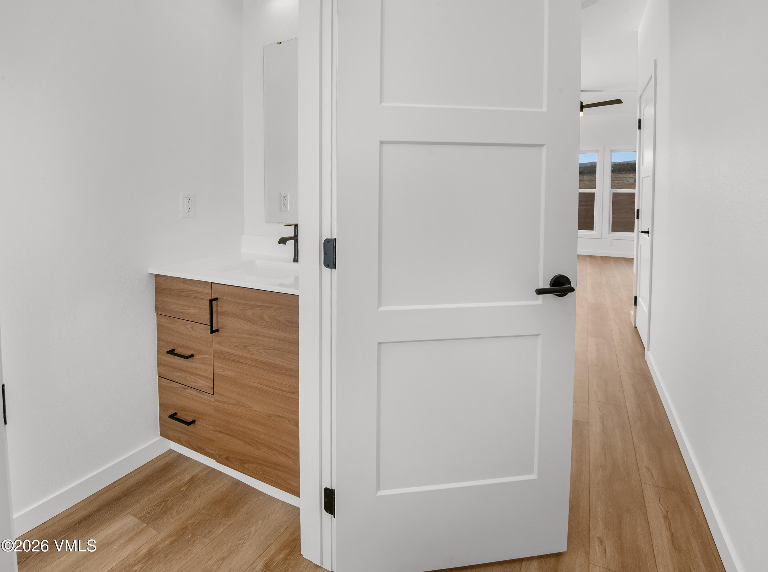 Bathroom vanity with wood-grain cabinetry beside an open door to a bright room.