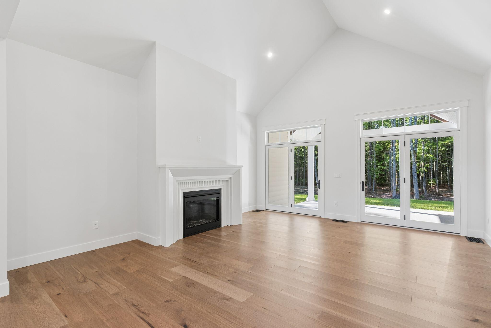 Vacant living room with a fireplace, vaulted ceiling, hardwood floors, and glass doors overlooking trees.