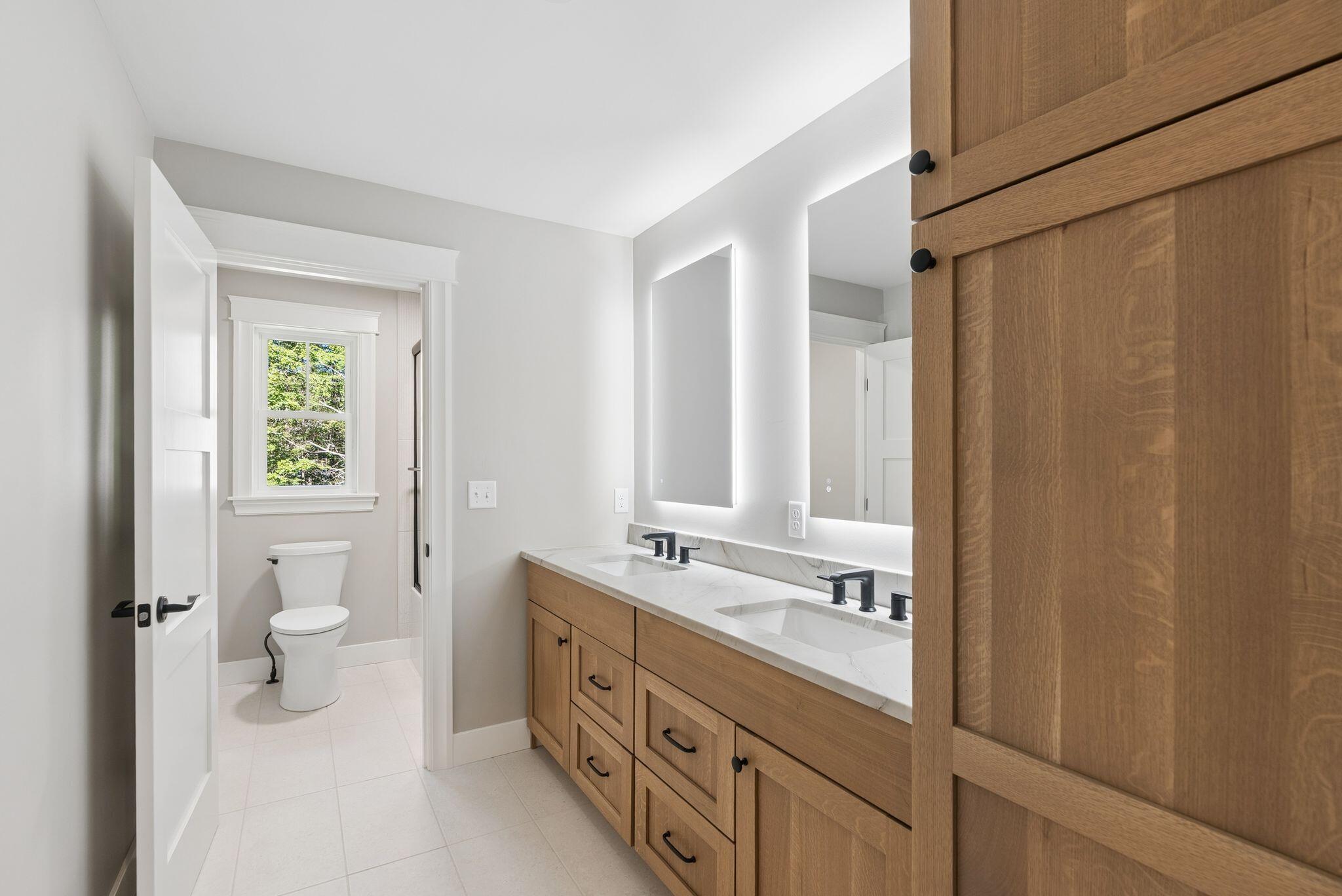 Bathroom with a double-sink wood vanity and a separate toilet area beside a window.