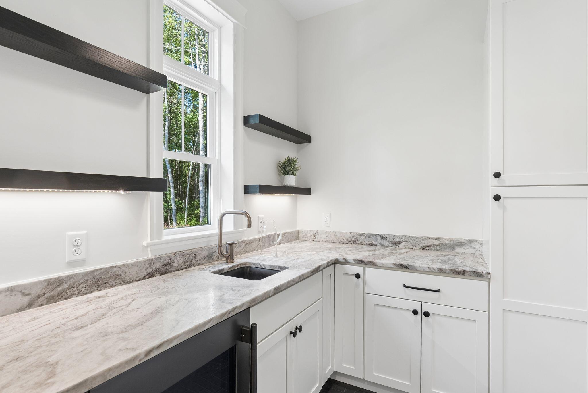 Kitchen with pale stone counters, a dark sink, floating shelves, and a tall window overlooking trees.