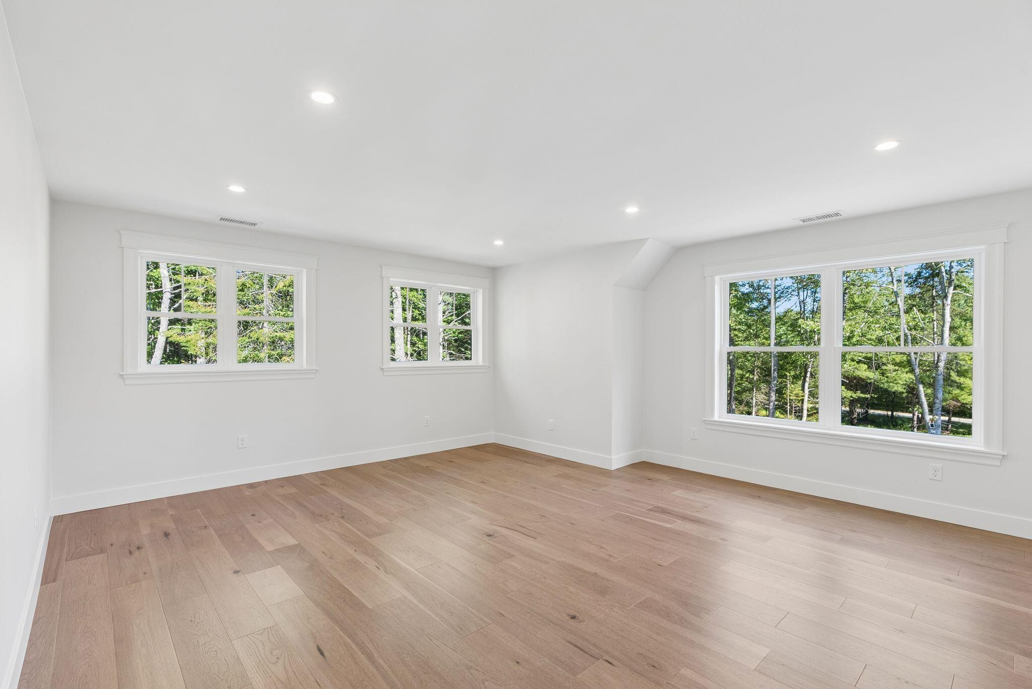 Empty bright room with wood floors, recessed lights, and windows overlooking trees.