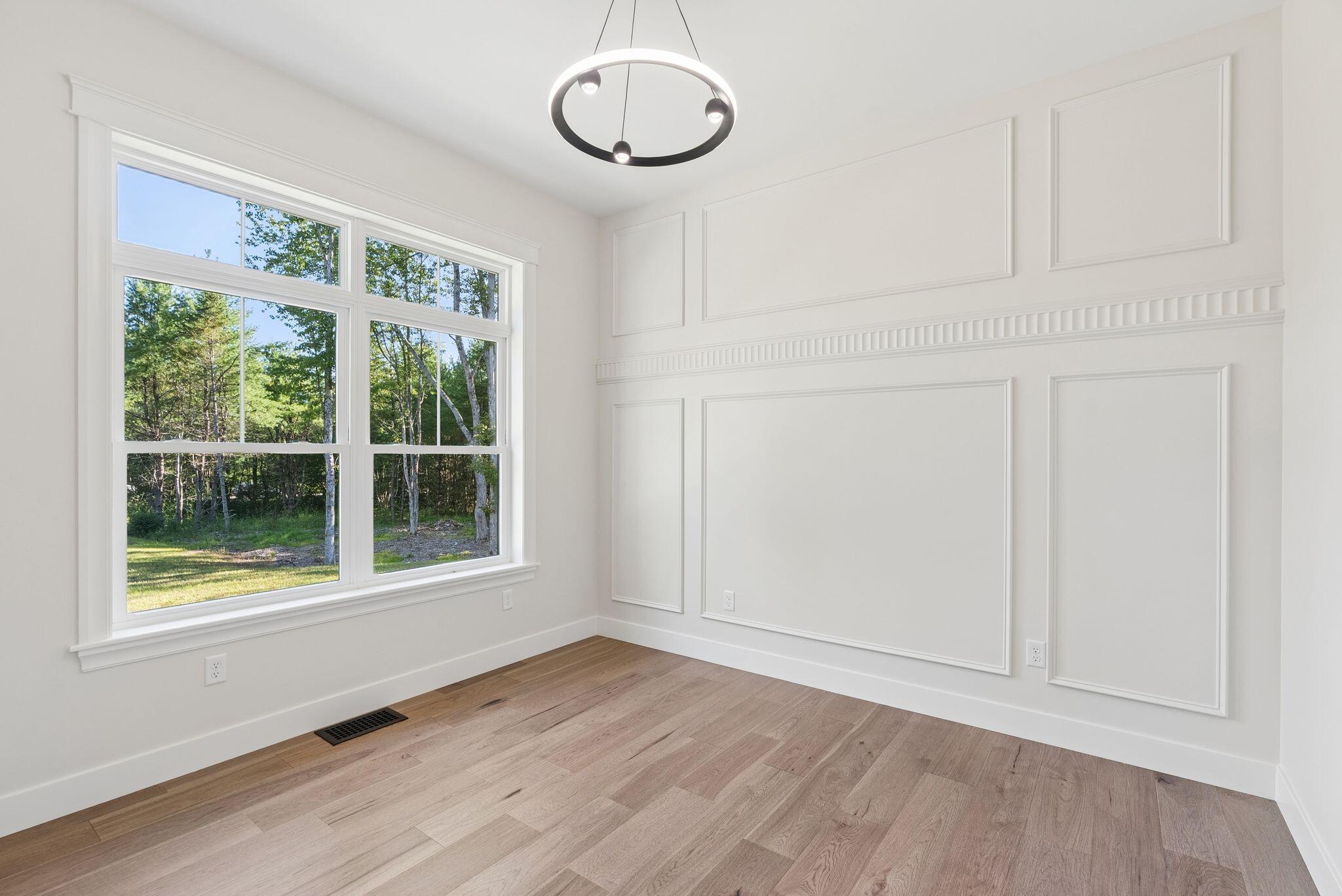 Vacant room with decorative wall paneling, wide-plank wood floors, and large windows overlooking trees.