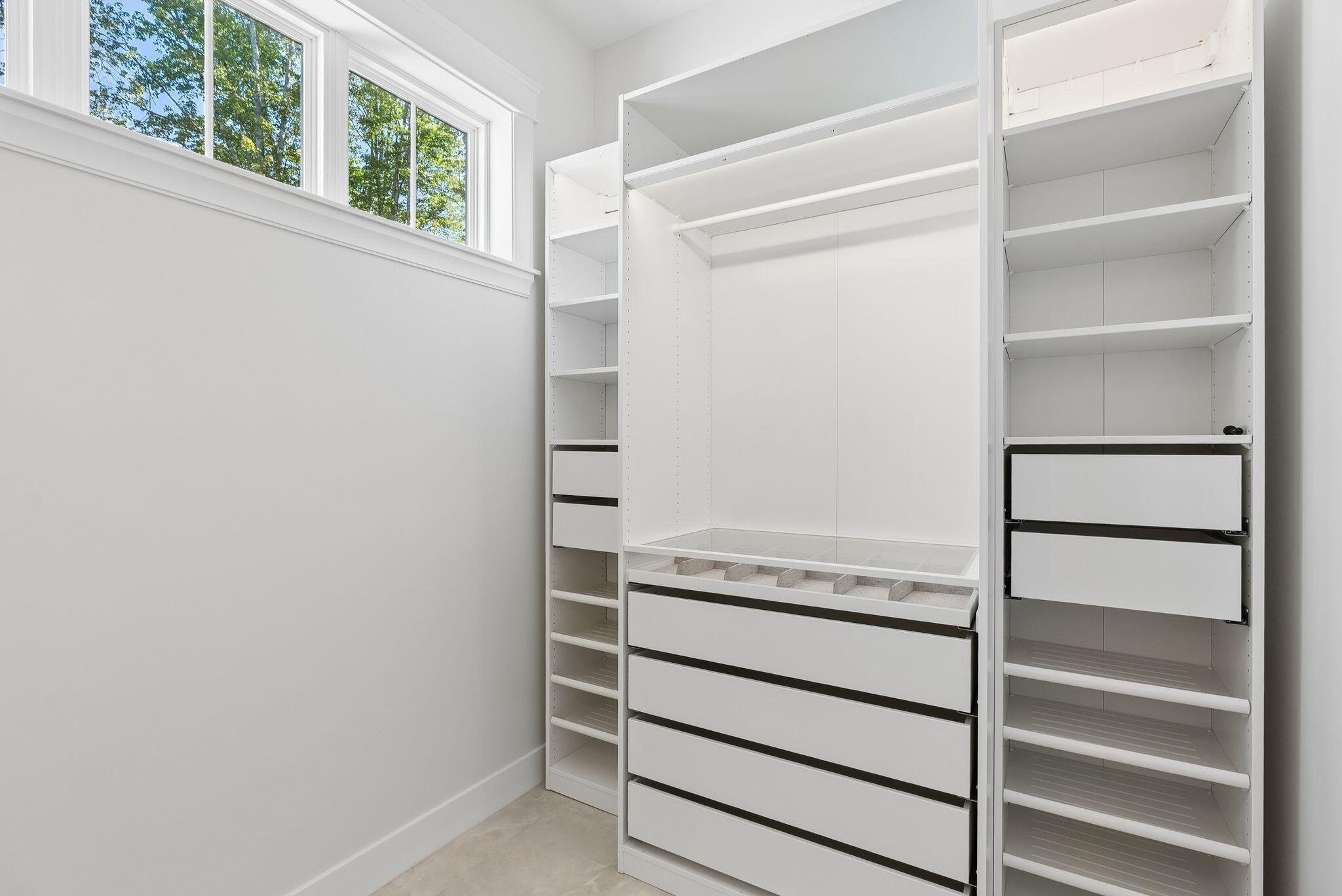 Walk-in closet with white built-in shelves and drawers beneath high windows.