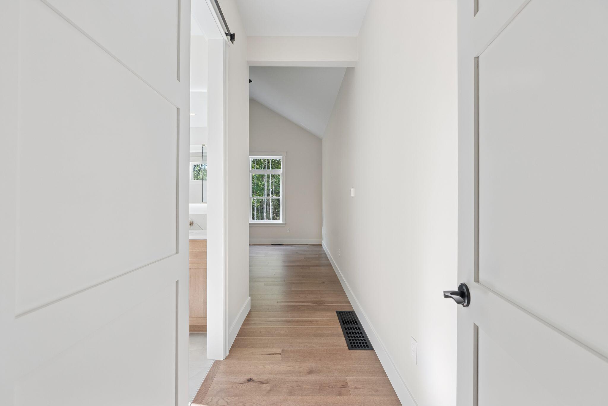 Bright hallway with wide-plank wood floors, a vaulted ceiling, and a window overlooking trees.