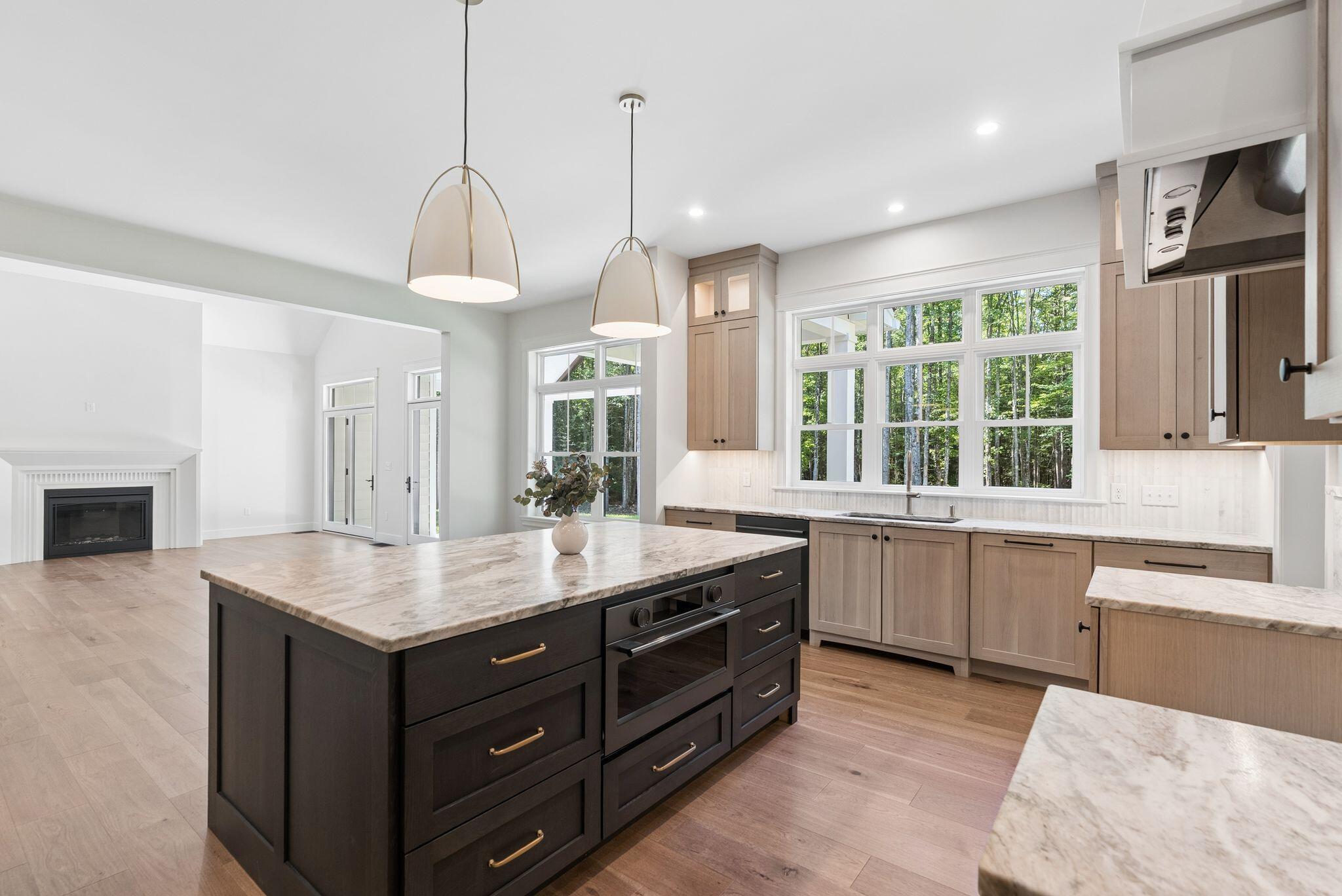 Open kitchen with a dark island, stone counters, light wood cabinets, and large windows facing trees.